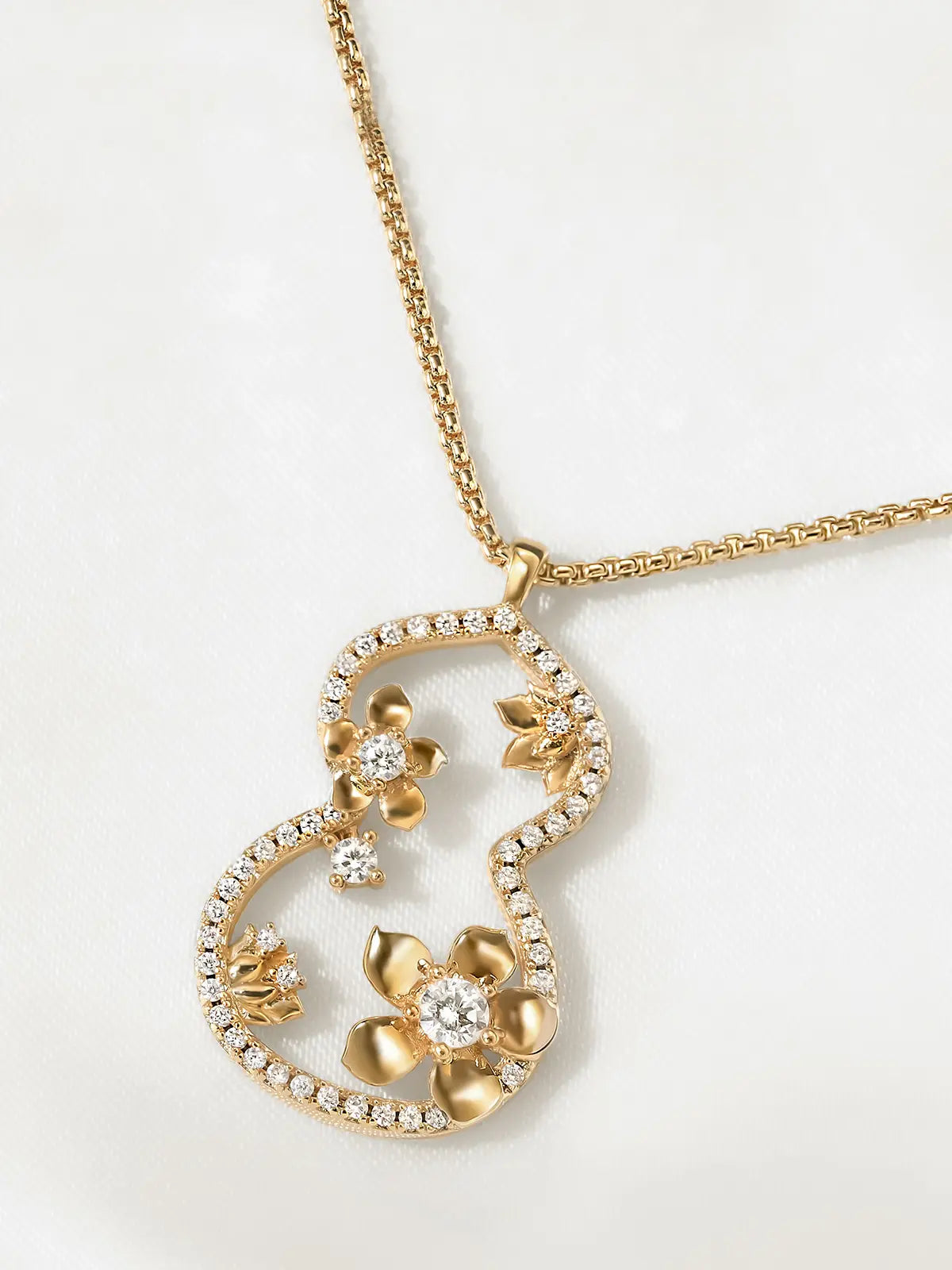 Close-up of the Tactilery Dream Gourd Window Necklace pendant on a white background, the large open-frame double-gourd silhouette with continuous pavé zircon inner border in detail — the sculptural interior garden scene showing two matte brushed gold five-petal flower blossoms with prong-set large round zircon centers, scattered small matte gold leaf sprigs, and two loose prong-set round zircon accent stones — with the polished gold box chain and bail visible above