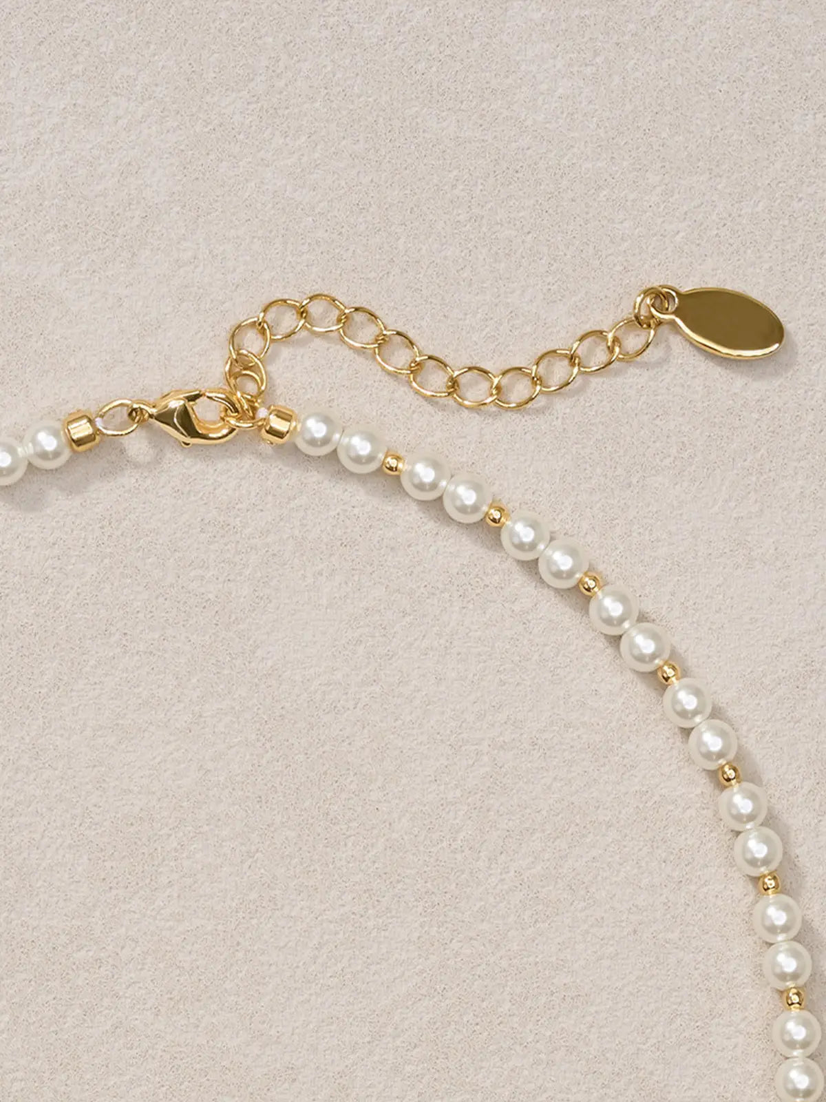 Close-up of the Tactilery Garden Floral Pearl Necklace clasp end on a warm cream surface, the polished gold lobster clasp and polished gold cable chain extension with oval tag charm visible, with the graduated freshwater pearl strand and small polished gold seed bead spacers shown in sharp detail — the alternating pearl and gold bead pattern clearly visible at the smaller-pearl clasp end