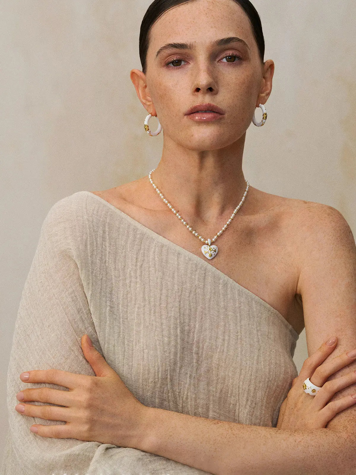 Three-quarter portrait of a model wearing the Tactilery Garden Floral Pearl Necklace with gaze downward, the graduated freshwater pearl strand with small polished gold seed bead spacers and central sculptural sunburst five-petal flower pendant with prong-set round zircon visible at the décolletage, against a warm taupe background wearing a white top with dark hair in a low bun