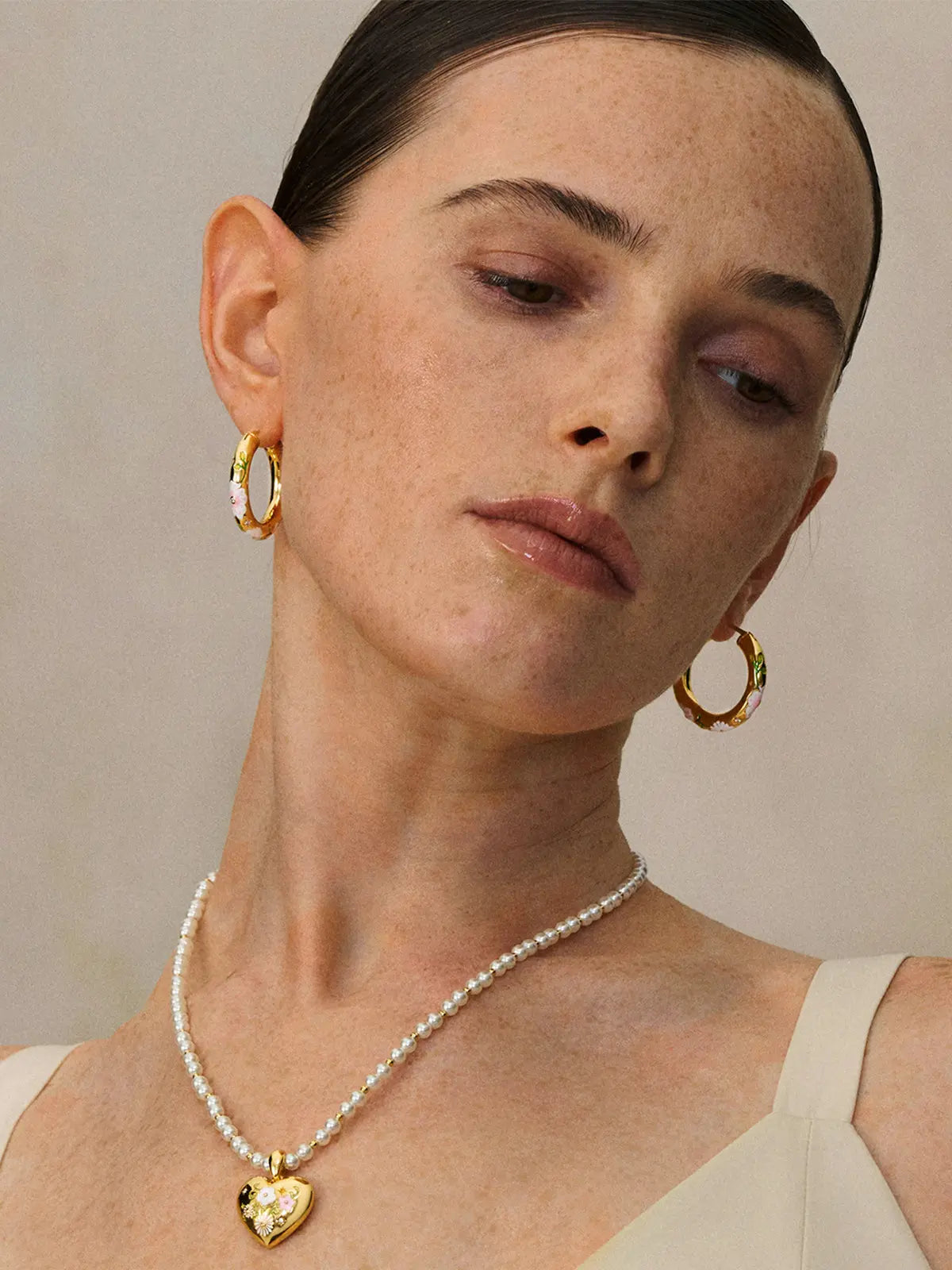 Close-up of a model's décolletage wearing the Tactilery Garden Floral Pearl Necklace from a slightly elevated angle, the graduated freshwater pearl strand with small polished gold seed bead spacers and central sculptural sunburst five-petal flower pendant with prong-set round zircon sitting at the neckline, against a warm taupe background wearing a white top