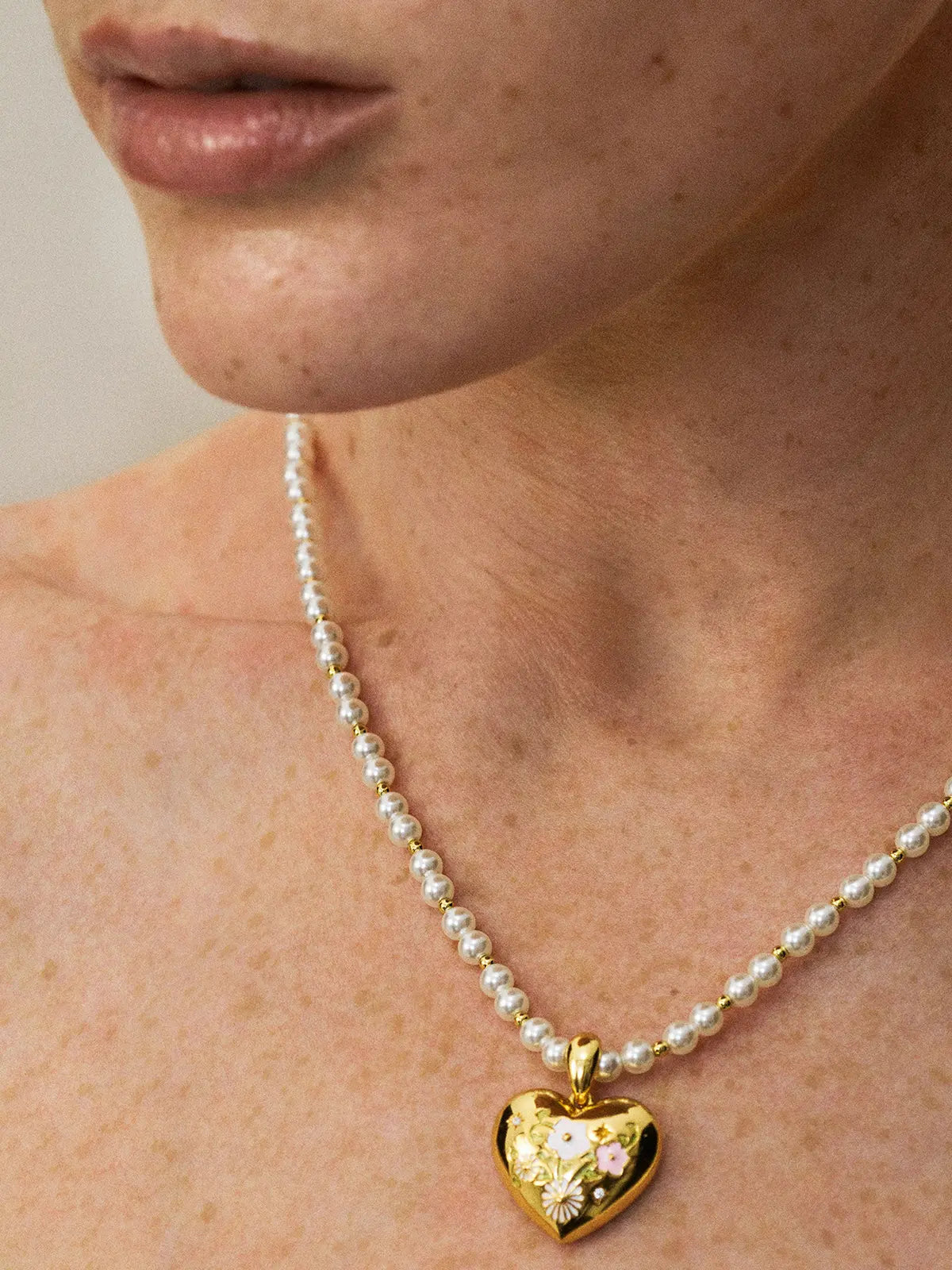 Three-quarter portrait of a model wearing the Tactilery Garden Floral Pearl Necklace with gaze to the side, the graduated freshwater pearl strand with small polished gold seed bead spacers and central sculptural sunburst five-petal flower pendant with prong-set round zircon visible at the décolletage, against a warm taupe background wearing a white top with dark hair pulled back
