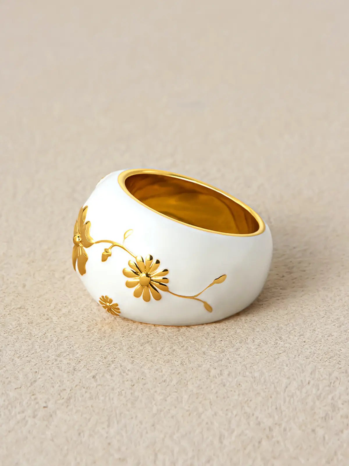 Tactilery Garden Relief Floral Ring displayed at an angle on a warm beige linen surface showing the three-dimensional depth of the wide white enamel dome band with raised gold botanical vine, daisy blooms, and leaf motifs, with polished gold interior visible