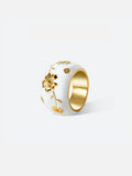 Tactilery Garden Relief Floral Ring displayed upright on a warm beige linen surface, the wide white enamel dome band with raised gold botanical vine, daisy blooms, and leaf motifs wrapping around the band, with polished gold interior visible at top