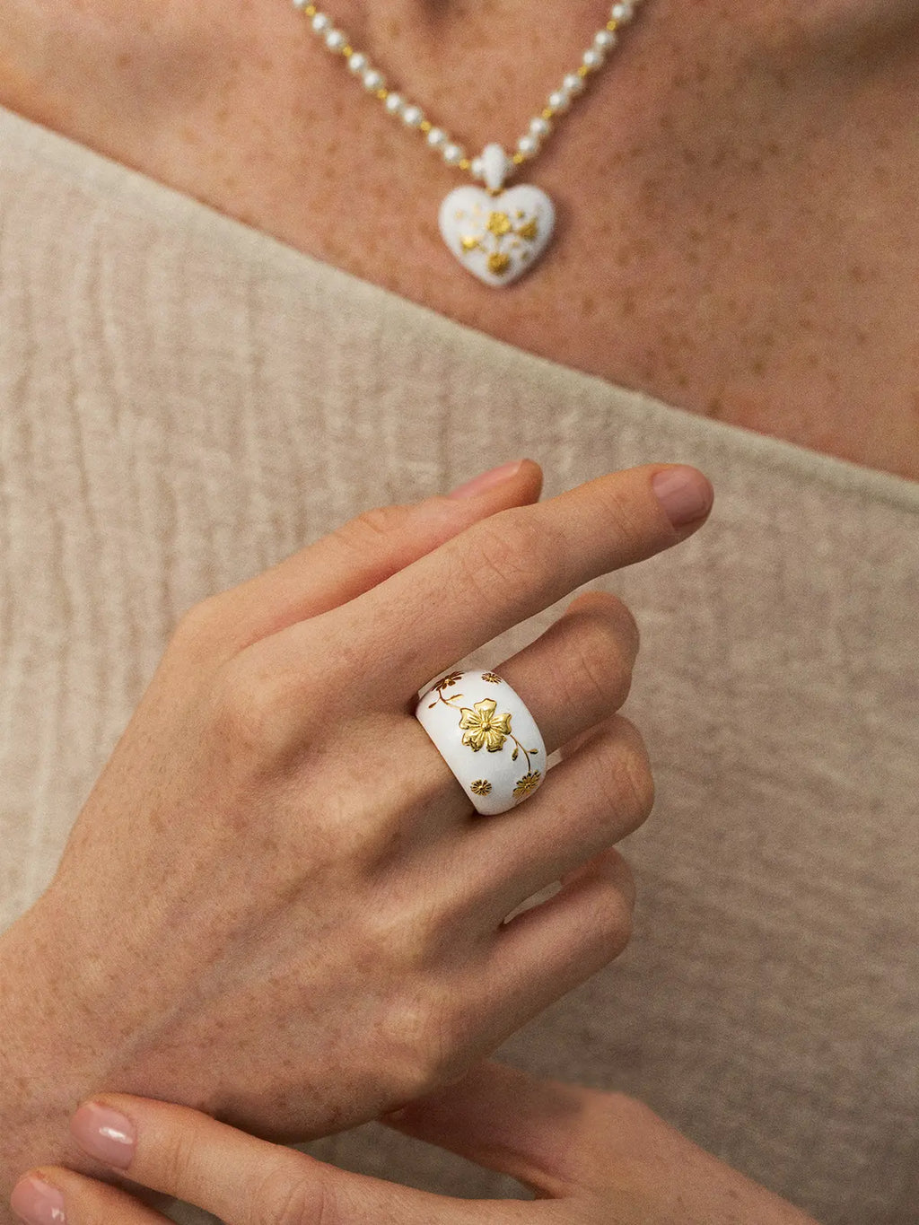 Model wearing the Tactilery Garden Relief Floral Ring on the middle finger, the wide white enamel dome band with raised gold daisy blooms and botanical vine motifs facing forward, paired with a white enamel heart pendant necklace on a pearl chain, against a warm beige background