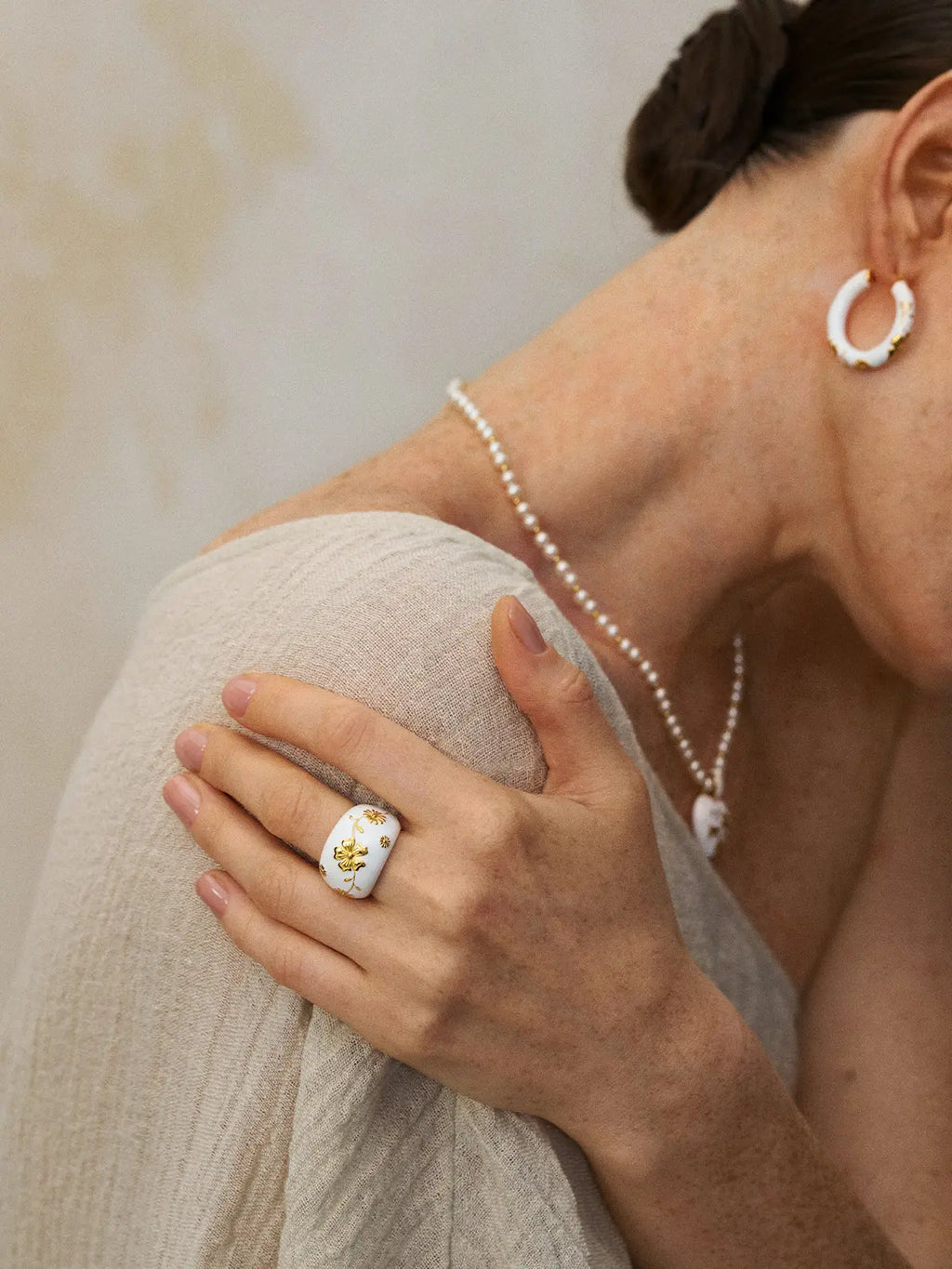 Editorial portrait of a model wearing the Tactilery Garden Relief Floral Ring on the index finger with hand raised to shoulder, the wide white enamel dome band with raised gold botanical vine and daisy motifs visible, paired with matching white enamel hoop earrings and a pearl chain necklace, against a warm grey background wearing a gold silk top