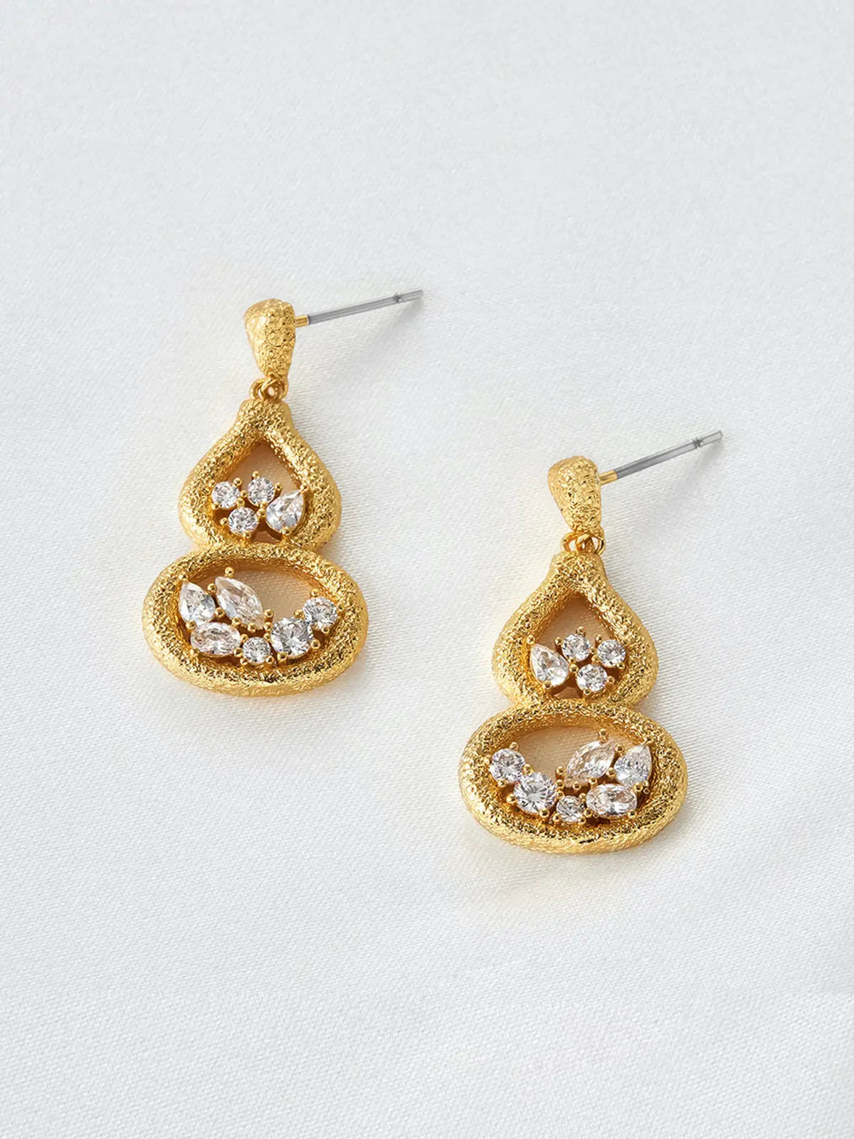 Pair of Tactilery Gourd Texture Stud Earrings in matte brushed gold displayed at an angle on a white surface, one showing the full front face of the double-gourd open frame with mixed-cut zircon clusters in each chamber, one showing the three-dimensional depth of the sand-textured gold frame, with silver post pins visible at top
