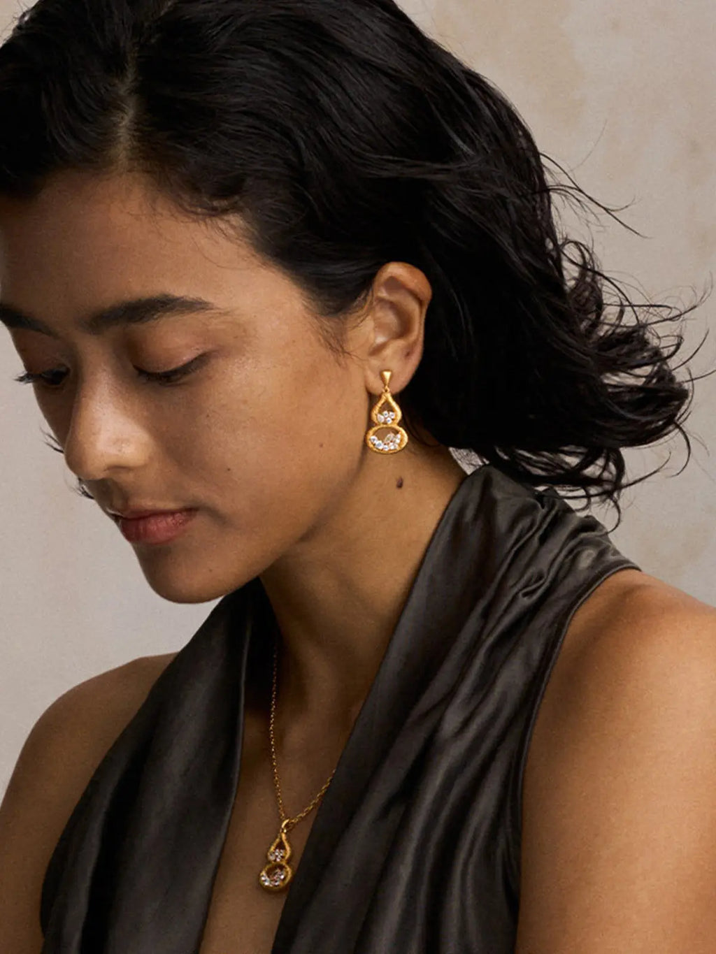 Three-quarter portrait of a model wearing the Tactilery Gourd Texture Stud Earrings with gaze downward, the double-gourd drop stud with heavily sand-textured gold open frame and mixed-cut zircon clusters dangling at the lobe, paired with a matching Gourd Texture pendant necklace on a fine gold chain, against a warm beige background wearing a dark charcoal draped sleeveless top