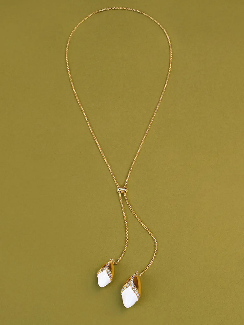 Tactilery Grain Rain Wheat Necklace in polished gold displayed laid out flat on an olive green surface showing the full necklace length, the fine polished gold cable chain forming a loop at top with the small rectangular slider bead at center and the two sculptural wheat grain pendants — smooth polished gold husk cap with V-shaped pavé zircon seam and smooth white ceramic grain body — hanging at the two chain ends at bottom