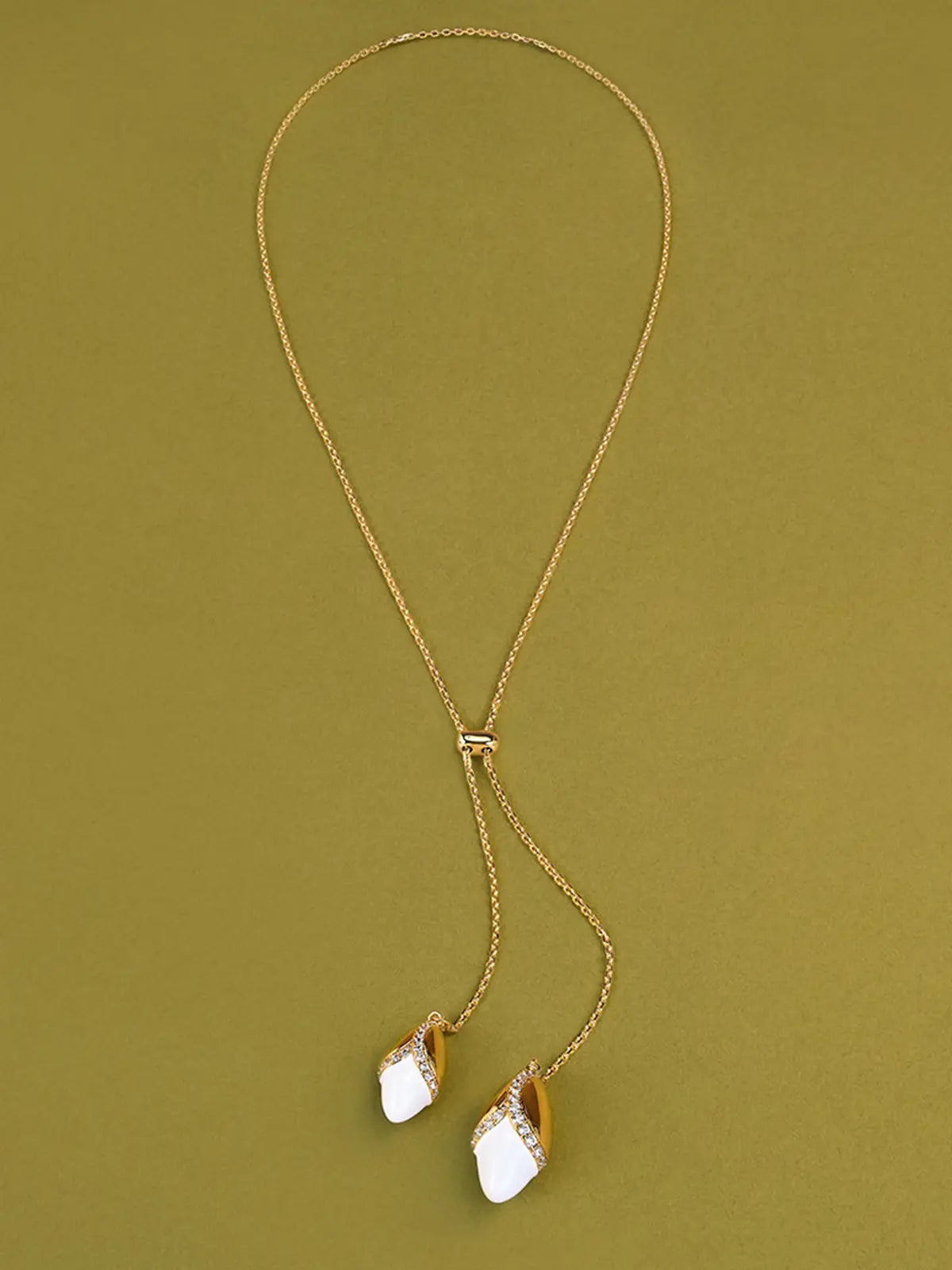 Tactilery Grain Rain Wheat Necklace in polished gold displayed laid out flat on an olive green surface showing the full necklace length, the fine polished gold cable chain forming a loop at top with the small rectangular slider bead at center and the two sculptural wheat grain pendants — smooth polished gold husk cap with V-shaped pavé zircon seam and smooth white ceramic grain body — hanging at the two chain ends at bottom