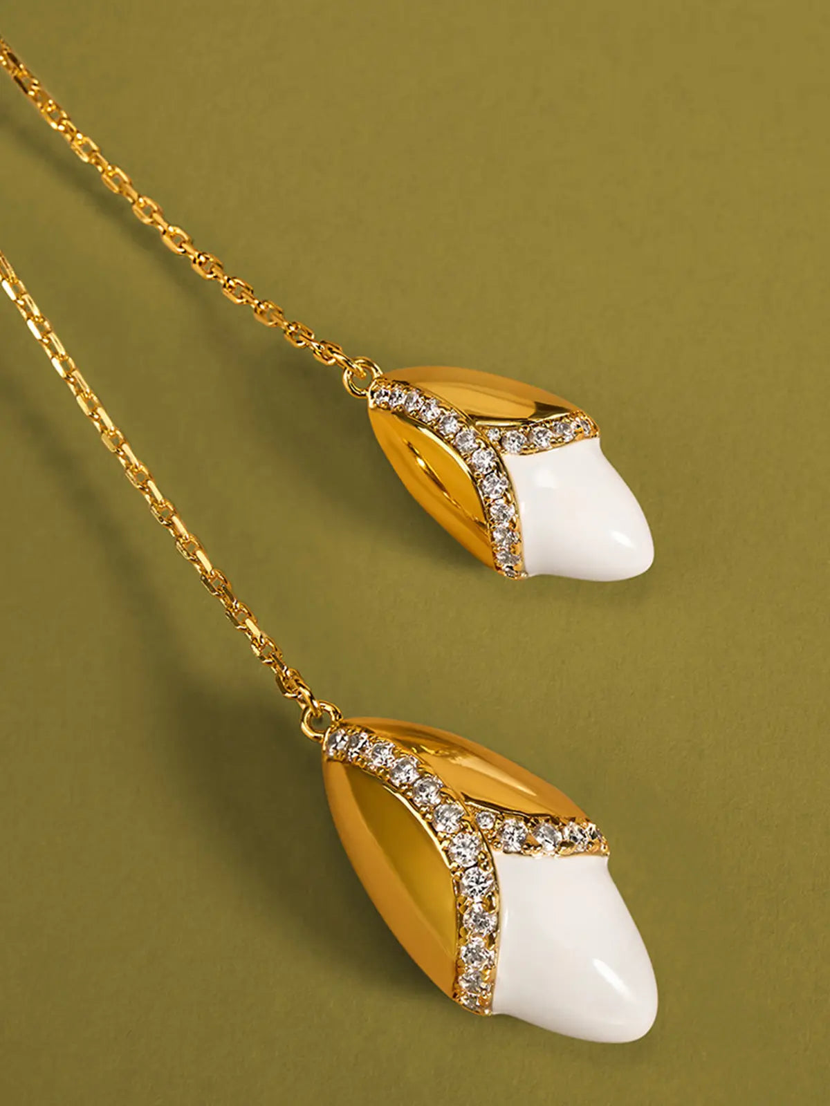Extreme close-up of the Tactilery Grain Rain Wheat Necklace two wheat grain pendants on an olive green surface, the two sculptural wheat grain pendants — smooth polished gold pointed oval husk cap with V-shaped pavé zircon seam line at the lower edge and smooth white ceramic pointed oval lower grain body — shown in sharp detail suspended from the fine polished gold cable chain ends