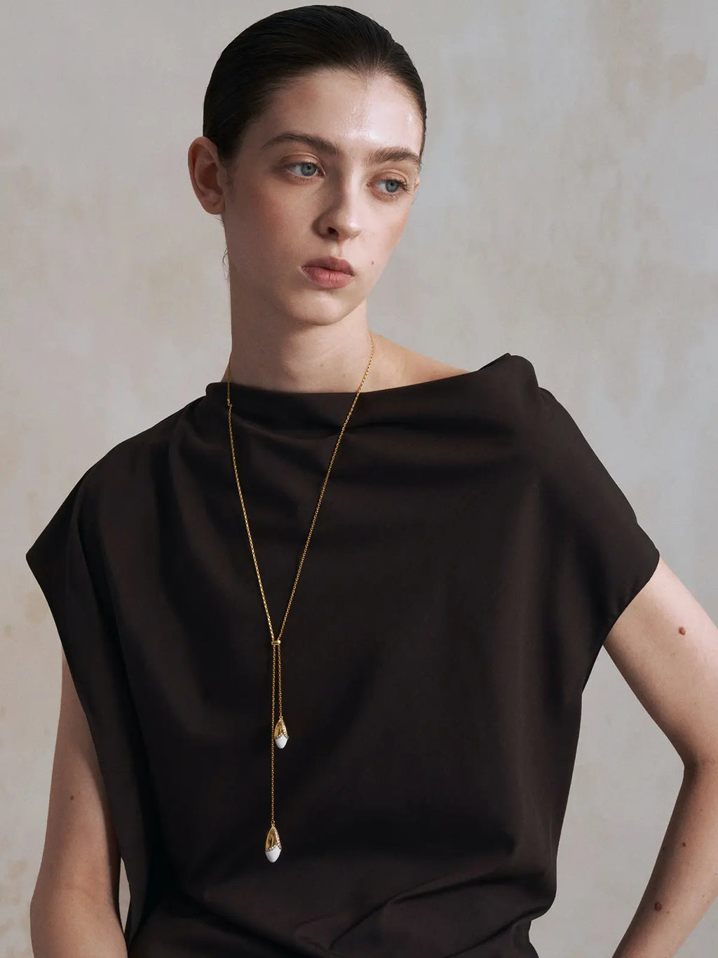 Front-facing editorial portrait of a model wearing the Tactilery Grain Rain Wheat Necklace with direct gaze, the adjustable lariat-style Y-drop with two sculptural wheat grain pendants — smooth polished gold husk cap with V-shaped pavé zircon seam and smooth white ceramic grain body — hanging at the sternum on a fine polished gold cable chain, against a warm grey background wearing a dark boat-neck top with dark hair slicked back