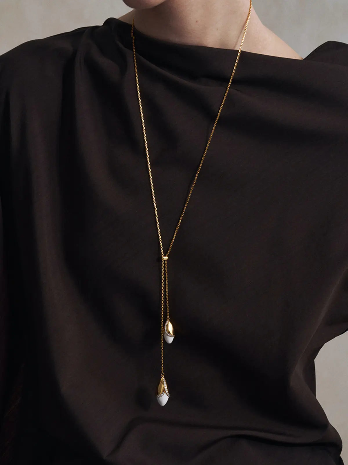 Close-up of a model's torso wearing the Tactilery Grain Rain Wheat Necklace, the adjustable lariat-style Y-drop with two sculptural wheat grain pendants — smooth polished gold husk cap with V-shaped pavé zircon seam and smooth white ceramic grain body — hanging at the sternum on a fine polished gold cable chain, against a warm grey background wearing a dark boat-neck top