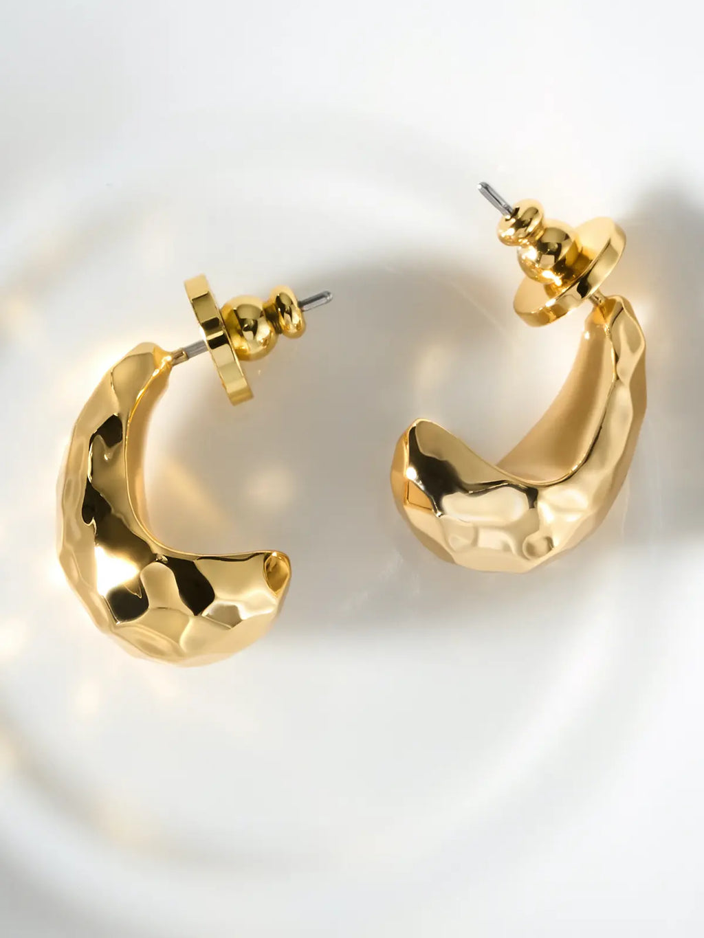 Pair of Tactilery Hammered Teardrop Earrings in polished gold displayed lying flat on a white ceramic dish, both showing the C-hoop profile with the wide chunky polished gold body and irregular hammered faceted surface, with gold ball screw-back fittings and silver post pins visible at top