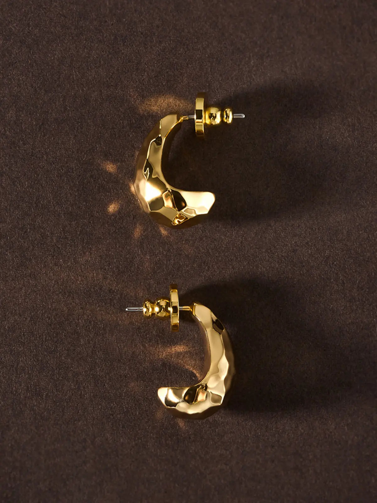 Pair of Tactilery Hammered Teardrop Earrings in polished gold displayed upright on a dark brown suede surface, both in C-hoop profile showing the wide chunky polished gold body with irregular hammered faceted surface, with gold ball screw-back fittings and silver post pins visible at top, casting dramatic shadows on the suede