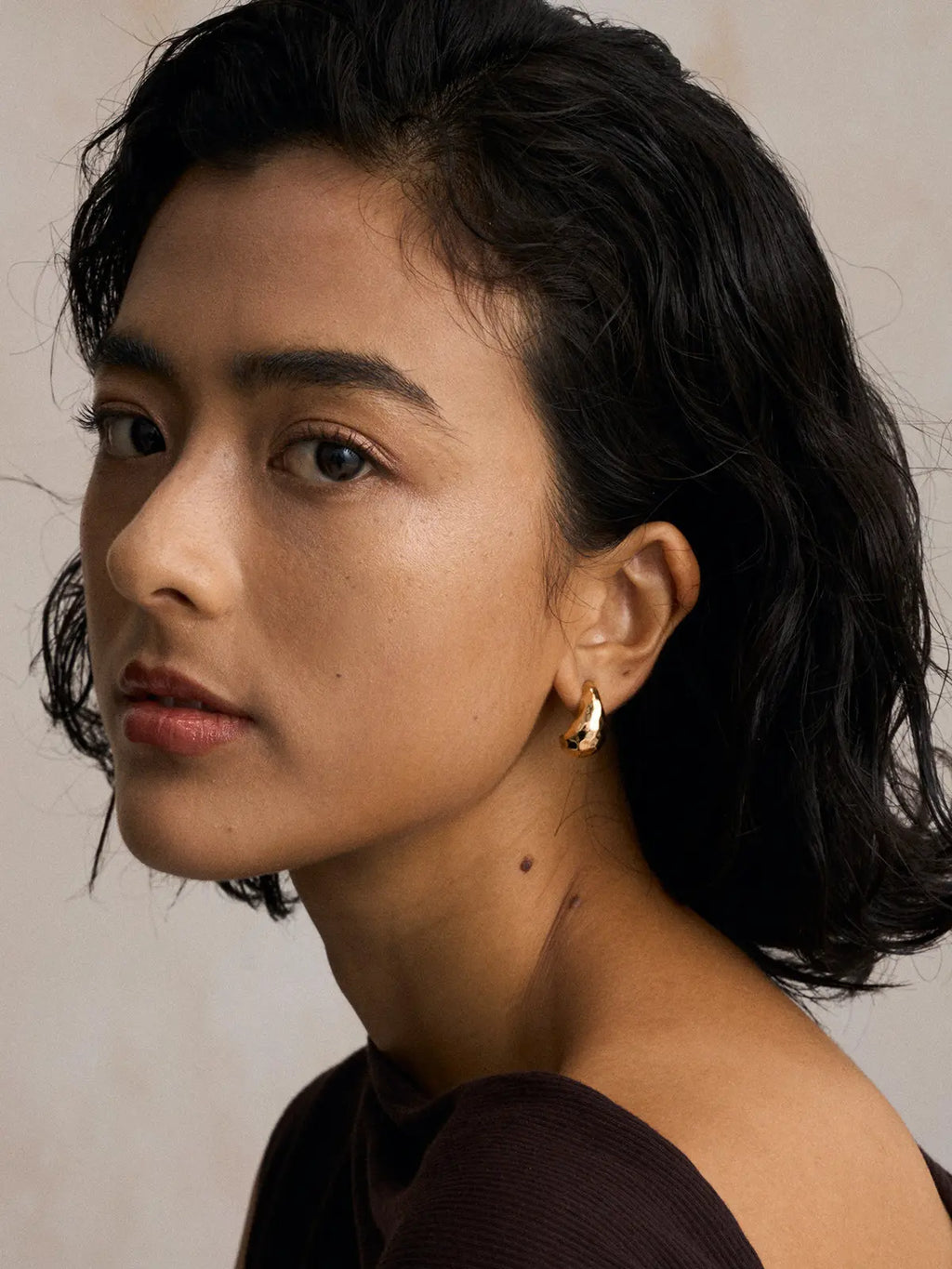 Three-quarter portrait of a model wearing the Tactilery Hammered Teardrop Earrings on both ears with gaze to the side, the wide chunky polished gold C-hoop with irregular hammered faceted surface sitting at the lobe, against a warm beige background wearing a dark brown long-sleeve top with short dark wavy hair