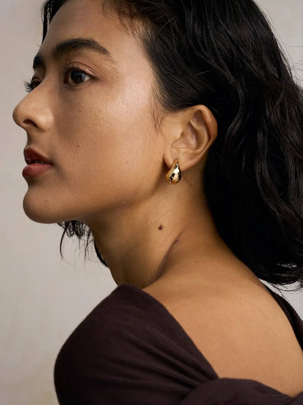 Side profile portrait of a model wearing the Tactilery Hammered Teardrop Earrings with gaze forward, the wide chunky polished gold C-hoop with irregular hammered faceted surface visible at the lobe, against a warm beige background wearing a dark brown long-sleeve top with short dark wavy hair