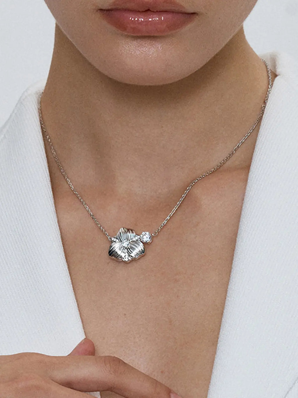Close-up of a model's décolletage wearing the Tactilery Hibiscus Flower Necklace, the three-dimensional matte brushed silver hibiscus flower pendant with four deeply ribbed petals, central prong-set large round zircon, pavé zircon petal seam accents, and floating chain zircon accent sitting at the neckline on a fine polished silver cable chain, against a cool grey background wearing a white structured blazer