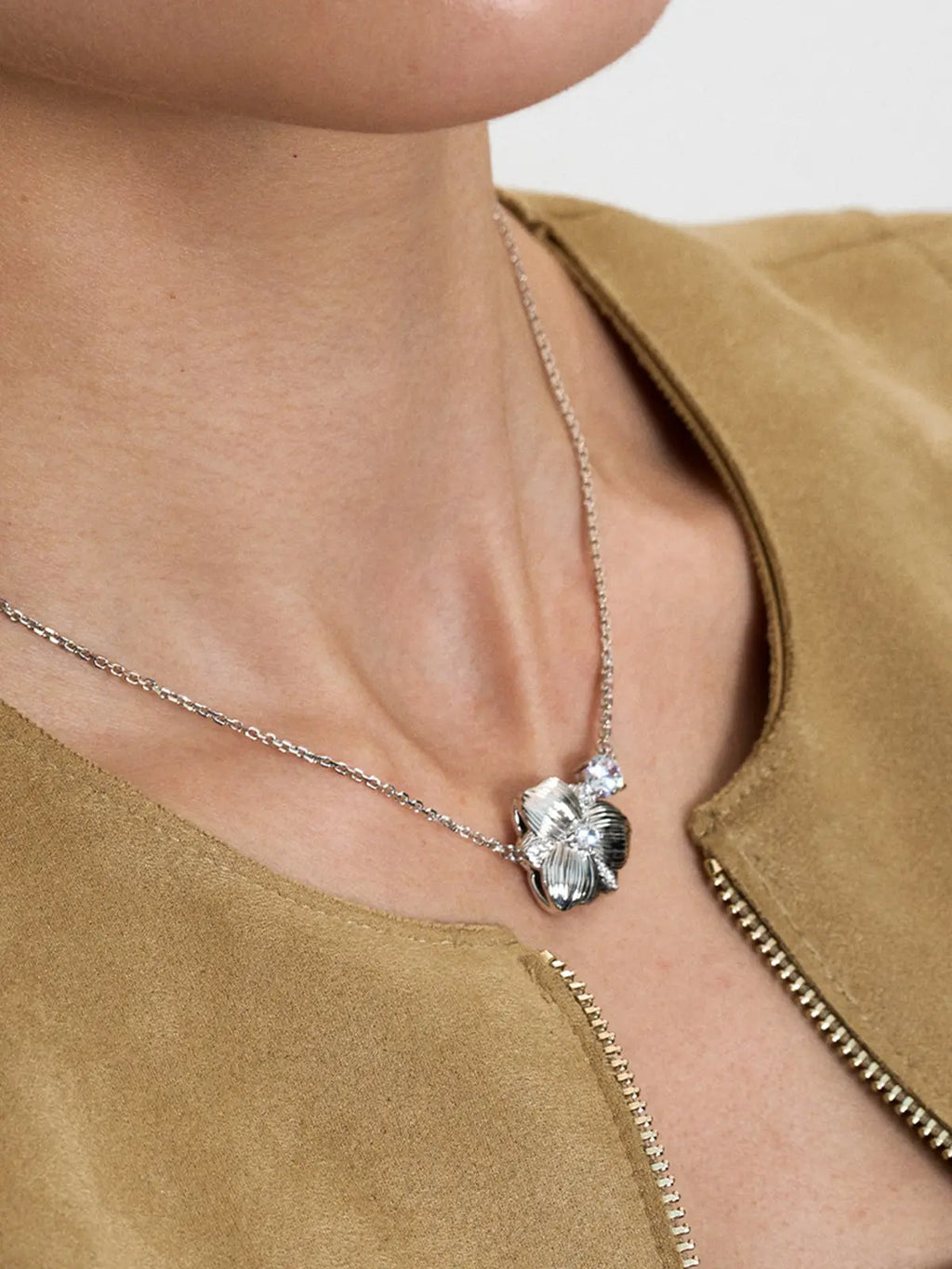 Close-up of a model's décolletage wearing the Tactilery Hibiscus Flower Necklace, the three-dimensional matte brushed silver hibiscus flower pendant with four deeply ribbed petals, central prong-set large round zircon, and floating chain zircon accent visible at the neckline on a fine polished silver cable chain, against a warm camel suede jacket background
