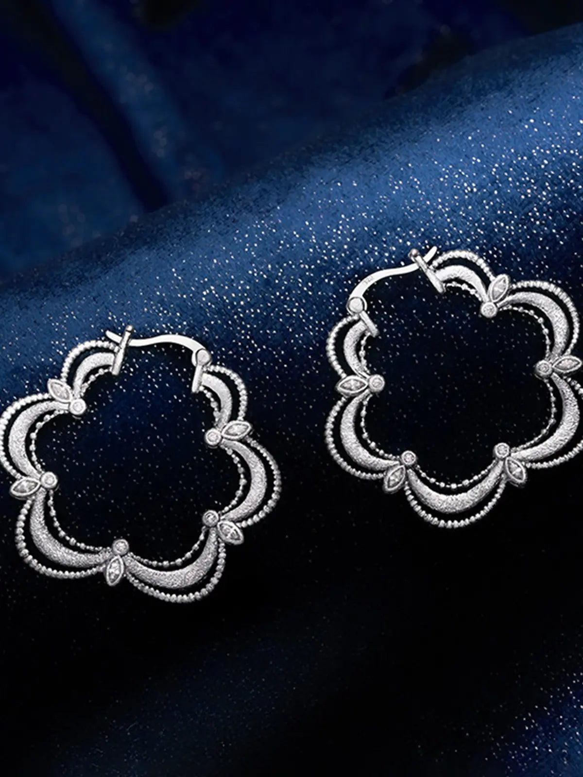 Pair of Tactilery Lace Court Earrings in polished silver displayed at an angle on a deep navy blue satin surface, both showing the three-dimensional depth of the quatrefoil hoop with scalloped four-lobe silhouette, three concentric polished silver wire arches per lobe with fine beaded milgrain borders, prong-set marquise zircon accents at each lobe junction, and small bezel-set round zircon at the base of each lobe, with hinged hoop post closures at top