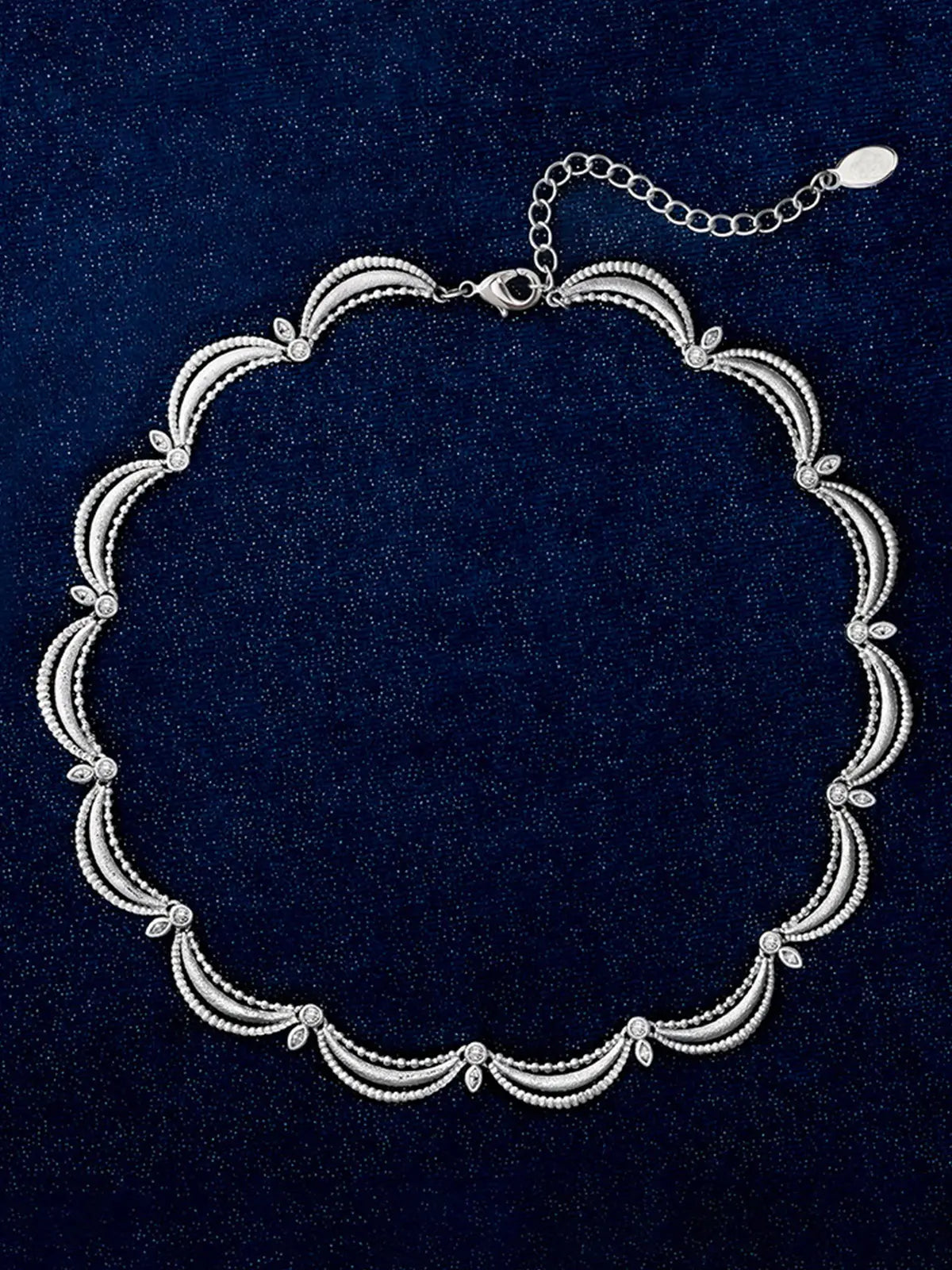 Tactilery Lace Court Necklace in polished silver displayed laid out flat on a dark navy velvet surface showing the full necklace length from above, the complete scalloped lace collar with all repeating arch link units, ball granulation bead edges, zircon junctions, and marquise zircon leaf accents visible, with the lobster clasp and oval tag charm extension chain at top right