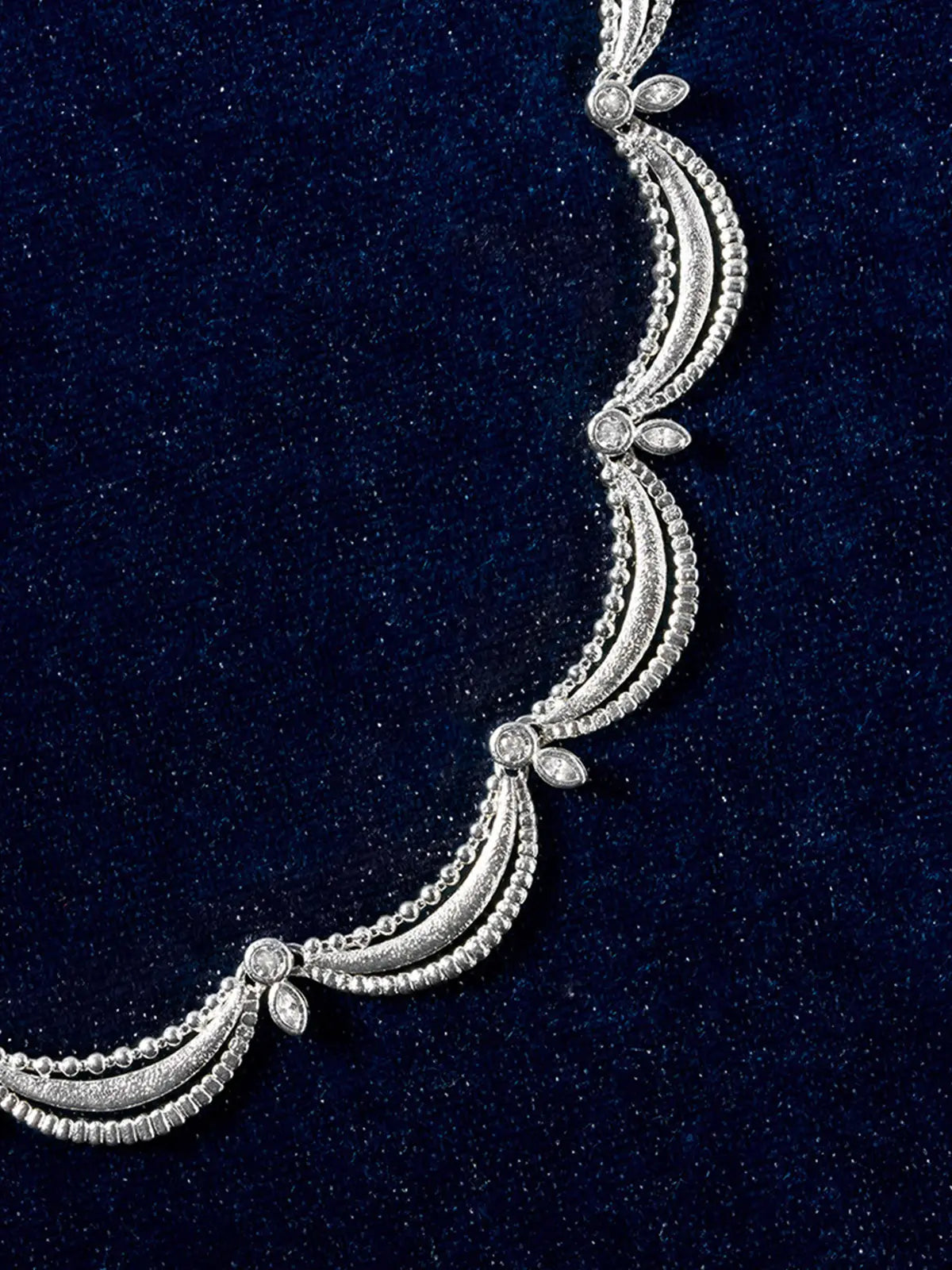Tactilery Lace Court Necklace in polished silver displayed laid out flat on a dark navy velvet surface showing the full necklace circle, the continuous row of repeating scalloped lace arch links with three concentric matte brushed silver crescent swag bands, ball granulation bead edges, bezel-set round zircon junctions, and marquise zircon leaf accents forming a complete scalloped lace collar circle, with the lobster clasp and oval tag charm extension chain visible at top