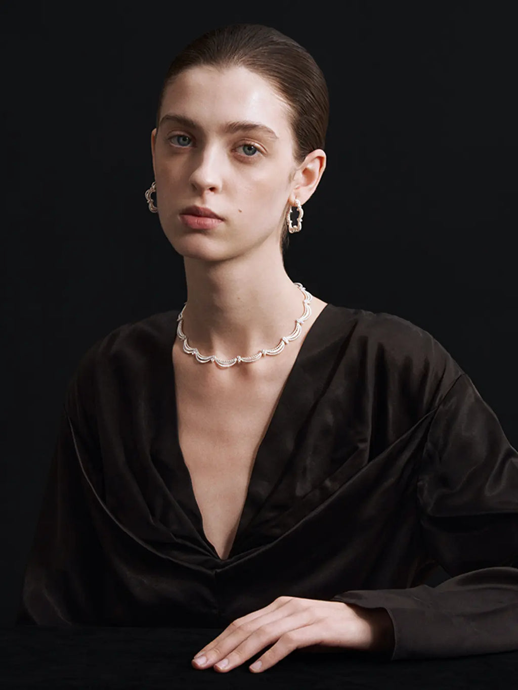 A model with slicked-back hair wearing a black satin draped top, a Tactilery lace court necklace, and matching silver hoop earrings, set against a solid black background.