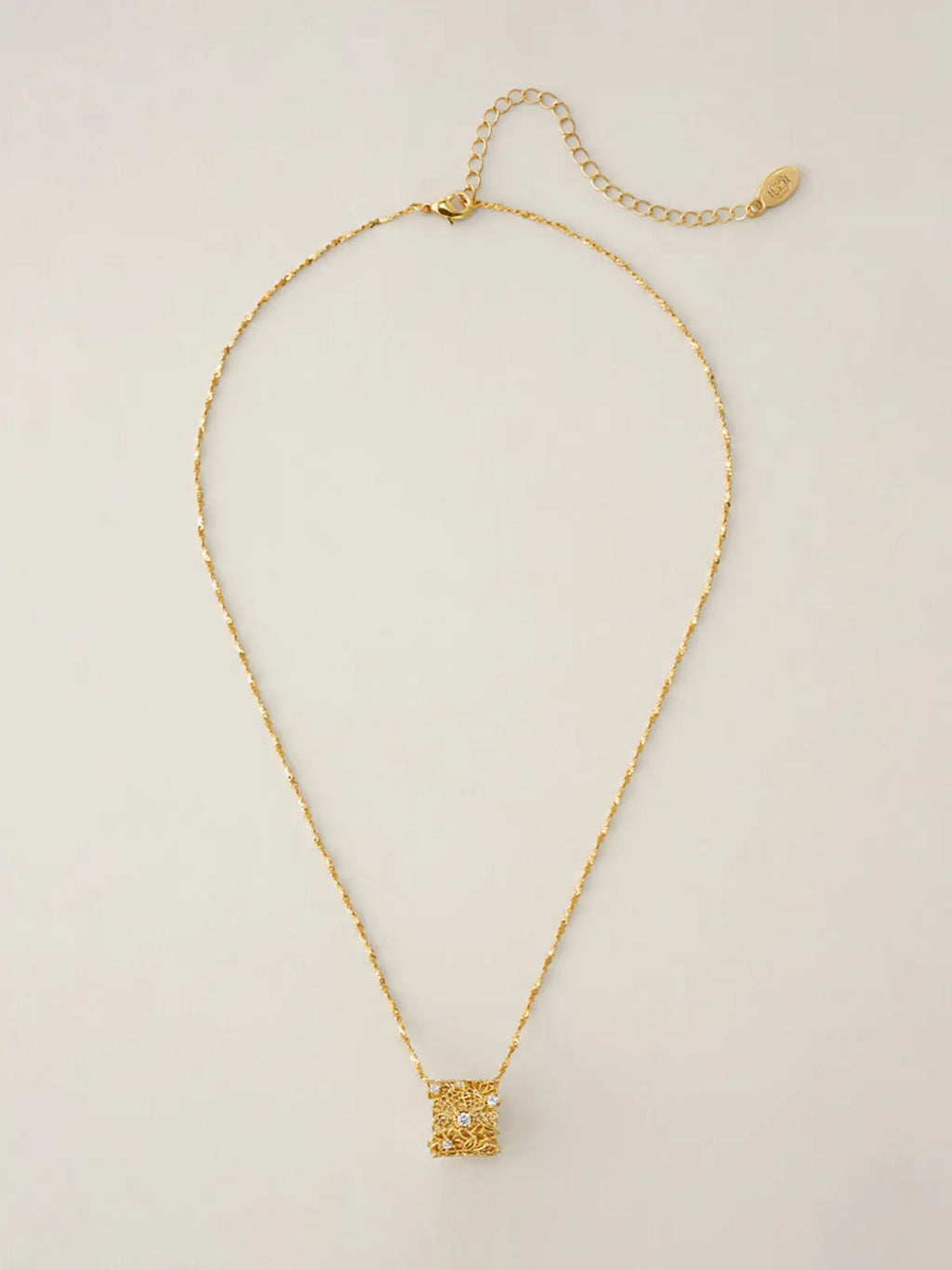 Tactilery Lace Garden Floating Necklace in polished gold displayed laid out flat on a warm cream surface showing the full necklace length, the fine polished gold twisted petal-link chain extending the full length with the lobster clasp and oval tag charm extension chain at top and the square openwork lace filigree pendant with scattered prong-set round zircons at bottom center