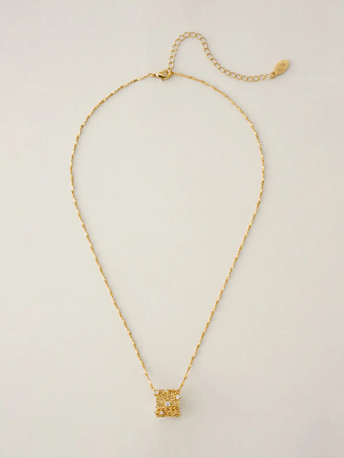 Tactilery Lace Garden Floating Necklace in polished gold displayed laid out flat on a warm cream surface showing the full necklace length, the fine polished gold twisted petal-link chain extending the full length with the lobster clasp and oval tag charm extension chain at top and the square openwork lace filigree pendant with scattered prong-set round zircons at bottom center