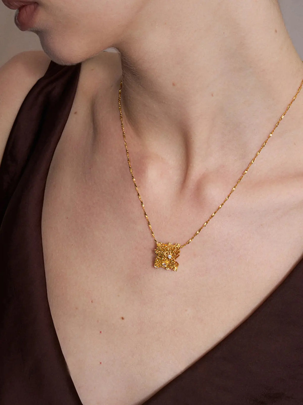 Close-up of a model's décolletage wearing the Tactilery Lace Garden Floating Necklace, the square openwork lace filigree gold pendant with scattered prong-set round zircons sitting at the neckline on a fine polished gold twisted petal-link chain, against a warm brown background wearing a dark draped top