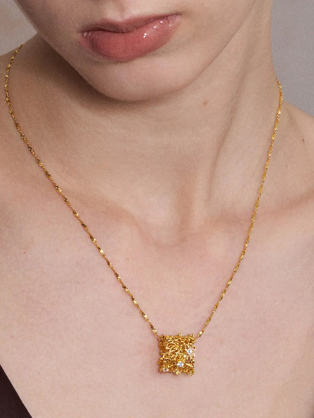 Close-up of a model's neck and décolletage wearing the Tactilery Lace Garden Floating Necklace from a slightly elevated angle, the square openwork lace filigree gold pendant with scattered prong-set round zircons hanging at the neckline on a fine polished gold twisted petal-link chain, against a warm taupe background wearing a dark draped top