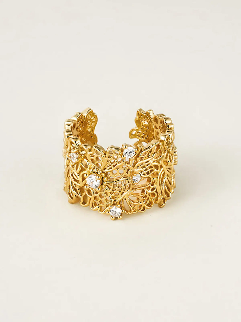 Tactilery Lace Garden Open Ring displayed upright on a warm beige surface showing the front face of the wide gold openwork lace floral cuff band with intricate vine and flower lattice pattern and five prong-set round zircon accents, open back visible at sides