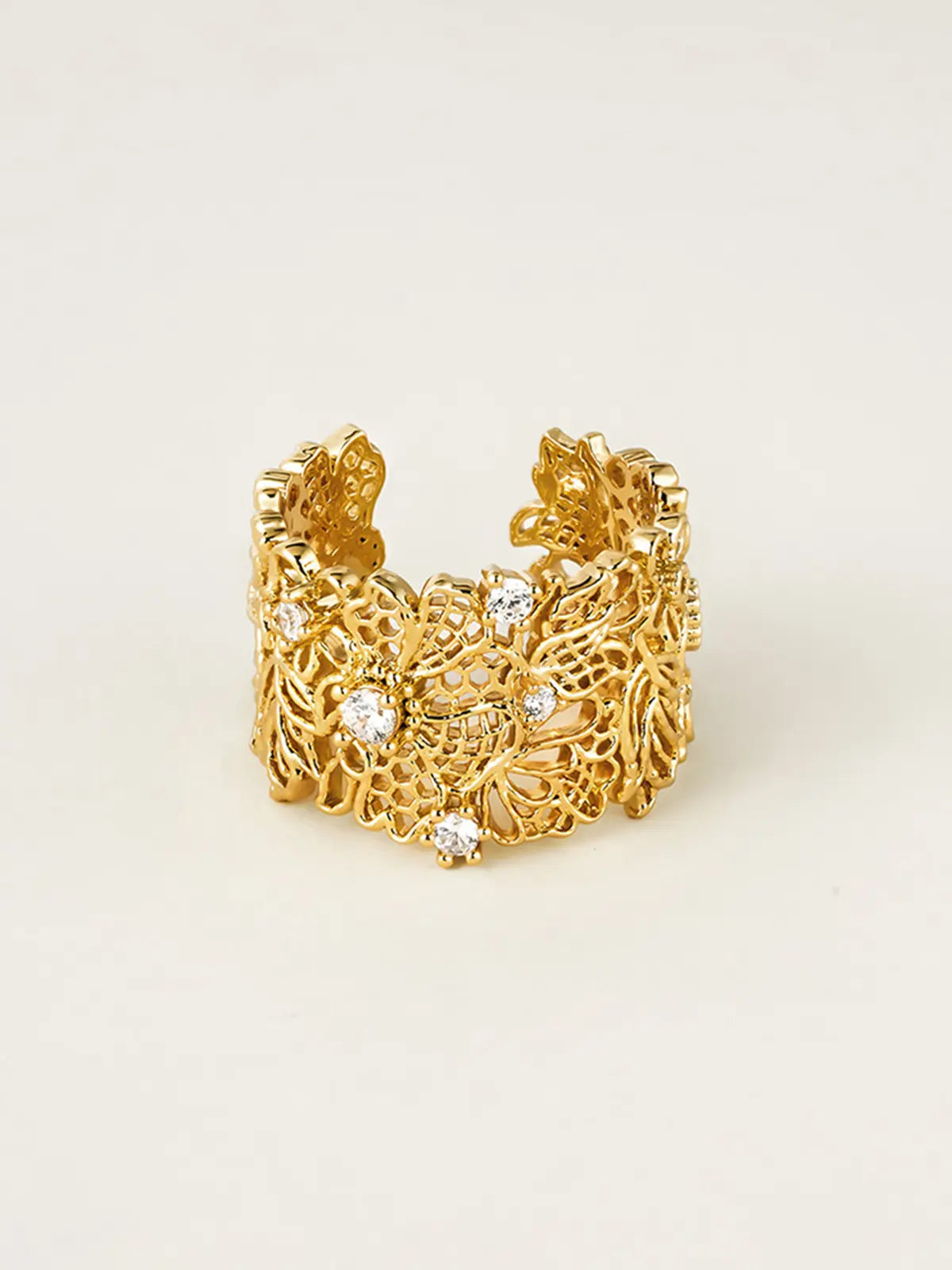 Tactilery Lace Garden Open Ring displayed upright on a warm beige surface showing the front face of the wide gold openwork lace floral cuff band with intricate vine and flower lattice pattern and five prong-set round zircon accents, open back visible at sides