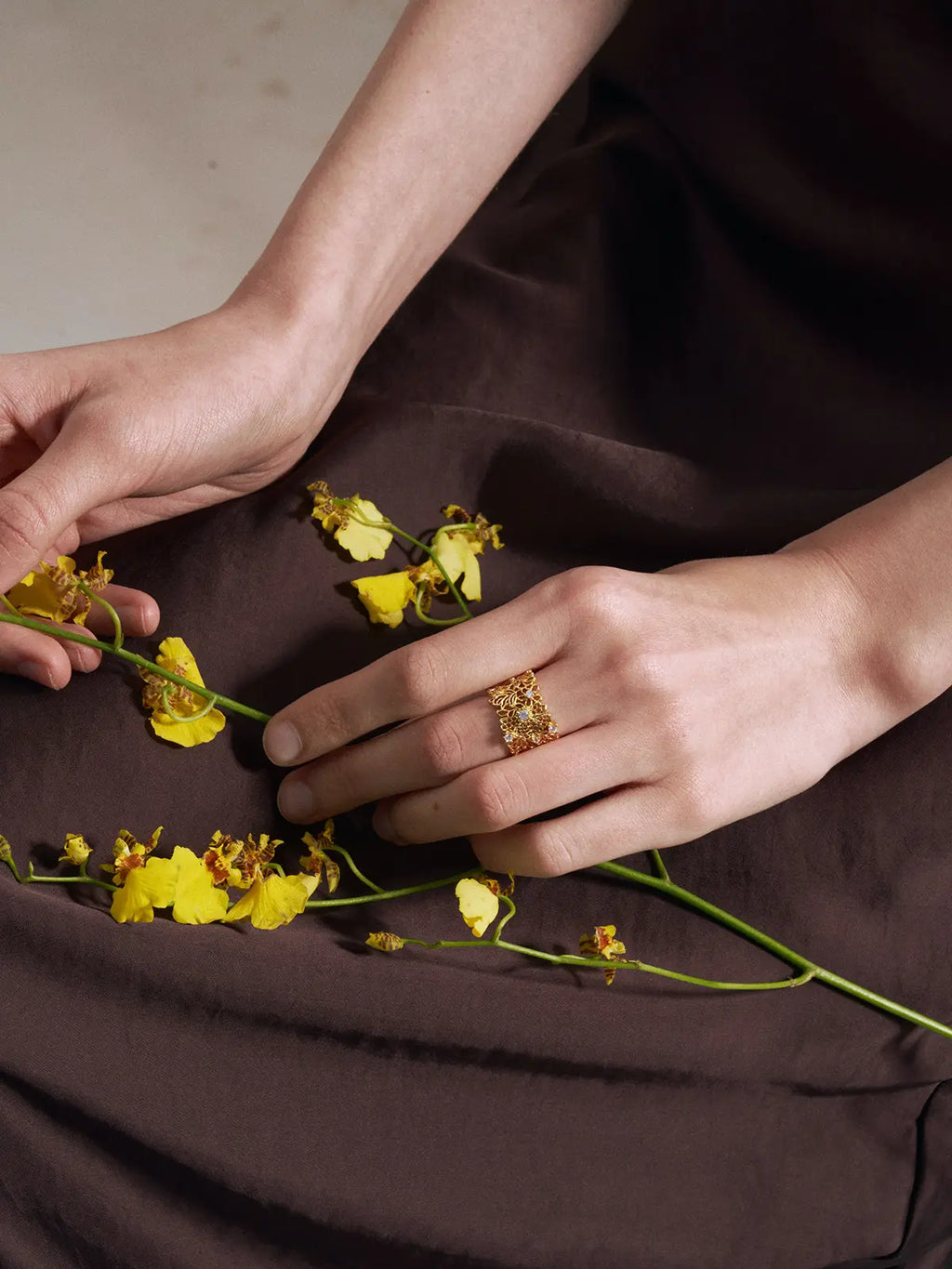 Editorial portrait of a model wearing the Tactilery Lace Garden Open Ring on the index finger with hand resting on shoulder, the wide gold openwork lace floral cuff band with five prong-set round zircon accents visible, paired with a delicate gold chain necklace, against a warm beige background