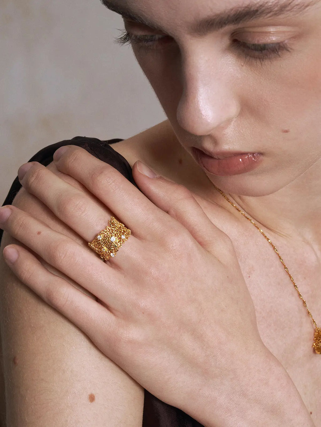 Model wearing the Tactilery Lace Garden Open Ring on the index finger with hand raised near face, the wide gold openwork lace floral cuff band with five prong-set round zircon accents visible, against a warm beige background