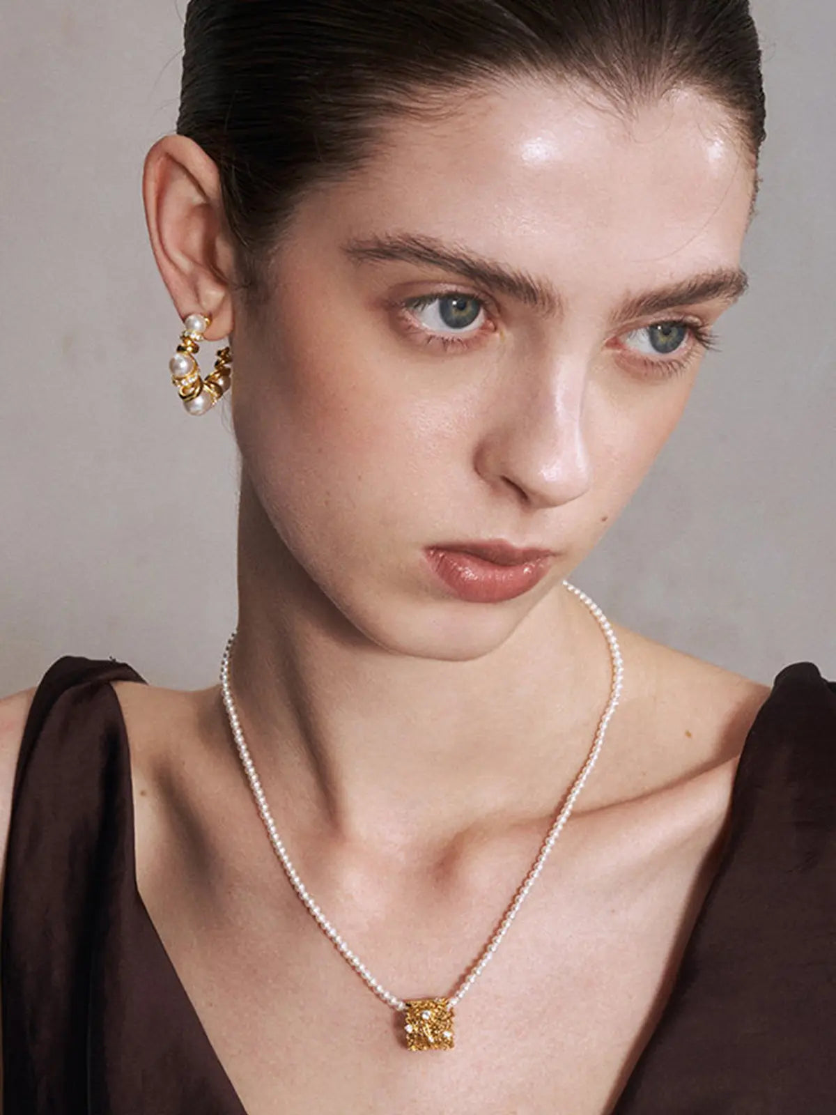 Close-up of a model's décolletage wearing the Tactilery Lace Garden Pearl Necklace from a slightly elevated angle, the square openwork lace filigree gold pendant with scattered prong-set round zircons and freshwater baroque pearl drop hanging at the neckline on a fine polished gold twisted petal-link chain, against a warm taupe background wearing a white draped top
