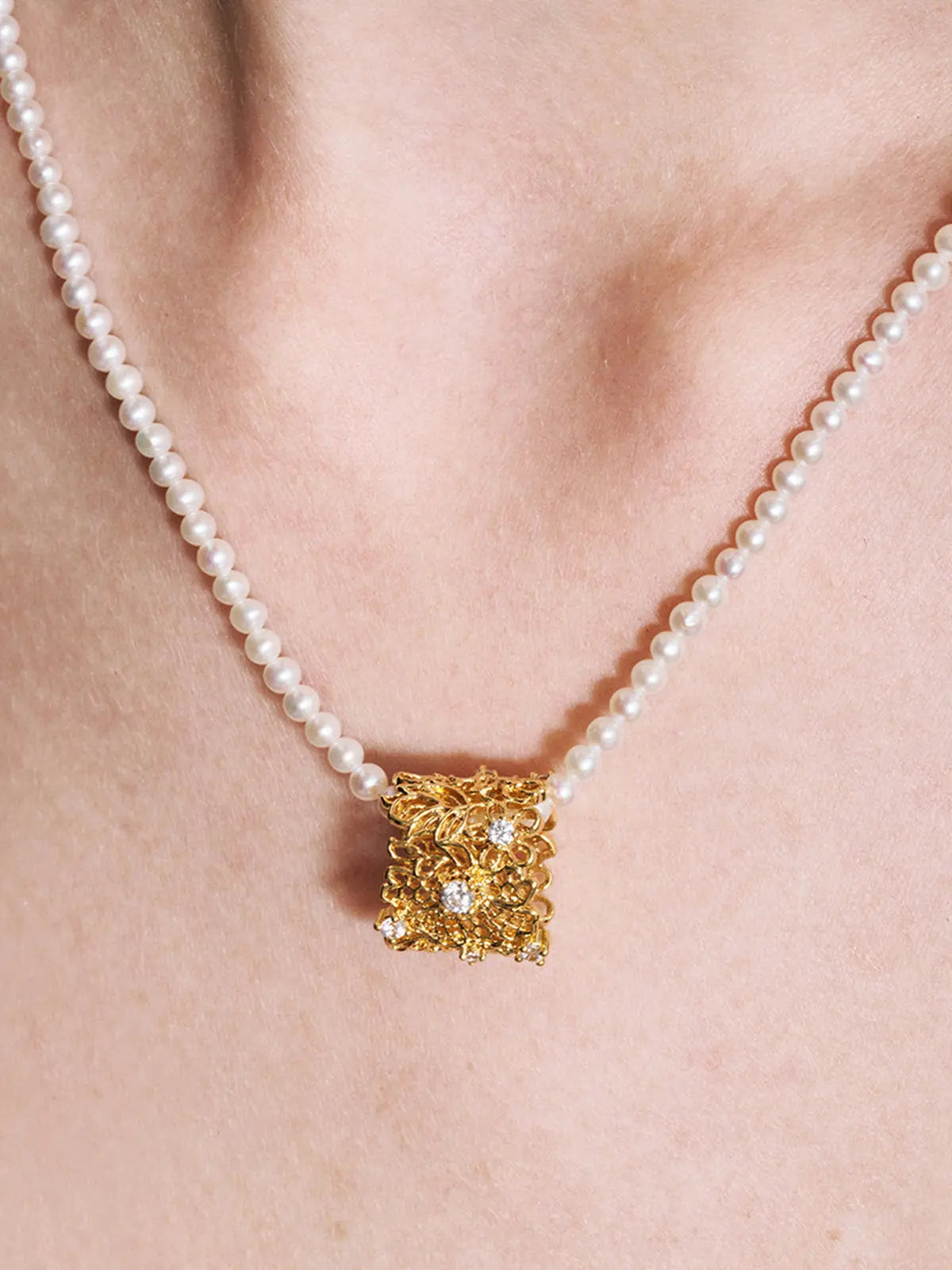 Three-quarter portrait of a model wearing the Tactilery Lace Garden Pearl Necklace with gaze downward, the square openwork lace filigree gold pendant with scattered prong-set round zircons and freshwater baroque pearl drop visible at the décolletage on a fine polished gold twisted petal-link chain, against a warm taupe background wearing a white draped top with dark hair in a low bun