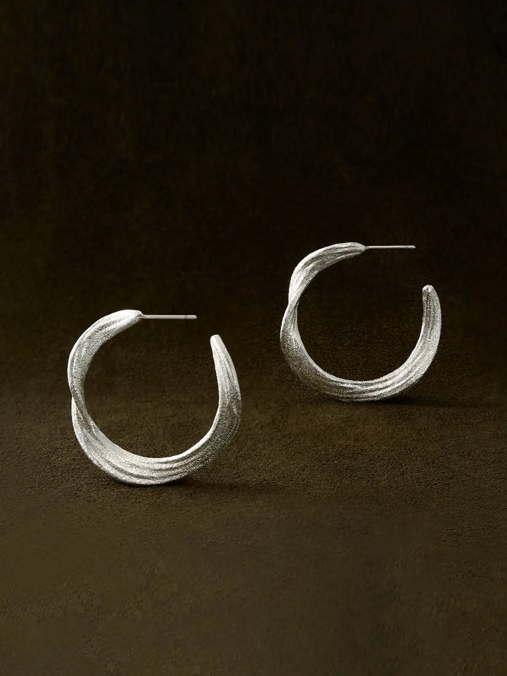 Pair of Tactilery Lake Ripple Hoop Earrings in matte brushed silver displayed upright on a dark brown suede surface, one face-on showing the full gently undulating wavy silhouette with fine horizontal brushed ripple texture, one in profile showing the three-dimensional depth of the wide flat band and the open C-hoop construction, with silver post pins visible at top