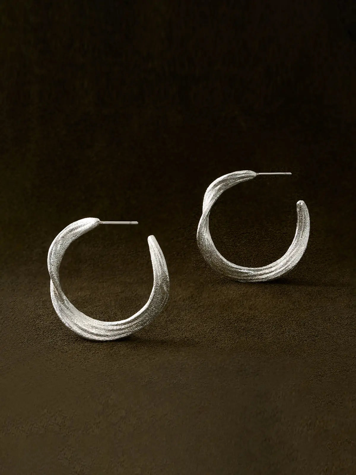Pair of Tactilery Lake Ripple Hoop Earrings in matte brushed silver displayed upright on a dark brown suede surface, one face-on showing the full gently undulating wavy silhouette with fine horizontal brushed ripple texture, one in profile showing the three-dimensional depth of the wide flat band and the open C-hoop construction, with silver post pins visible at top