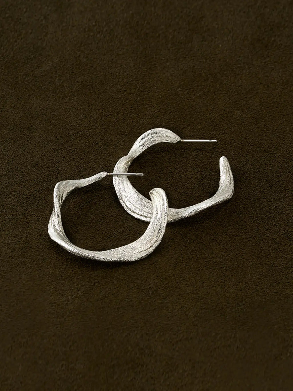 Pair of Tactilery Lake Ripple Hoop Earrings in matte brushed silver displayed lying flat and overlapping on a dark brown suede surface, showing the gently undulating wavy silhouette and fine horizontal brushed ripple texture across both hoops, with silver post pins visible