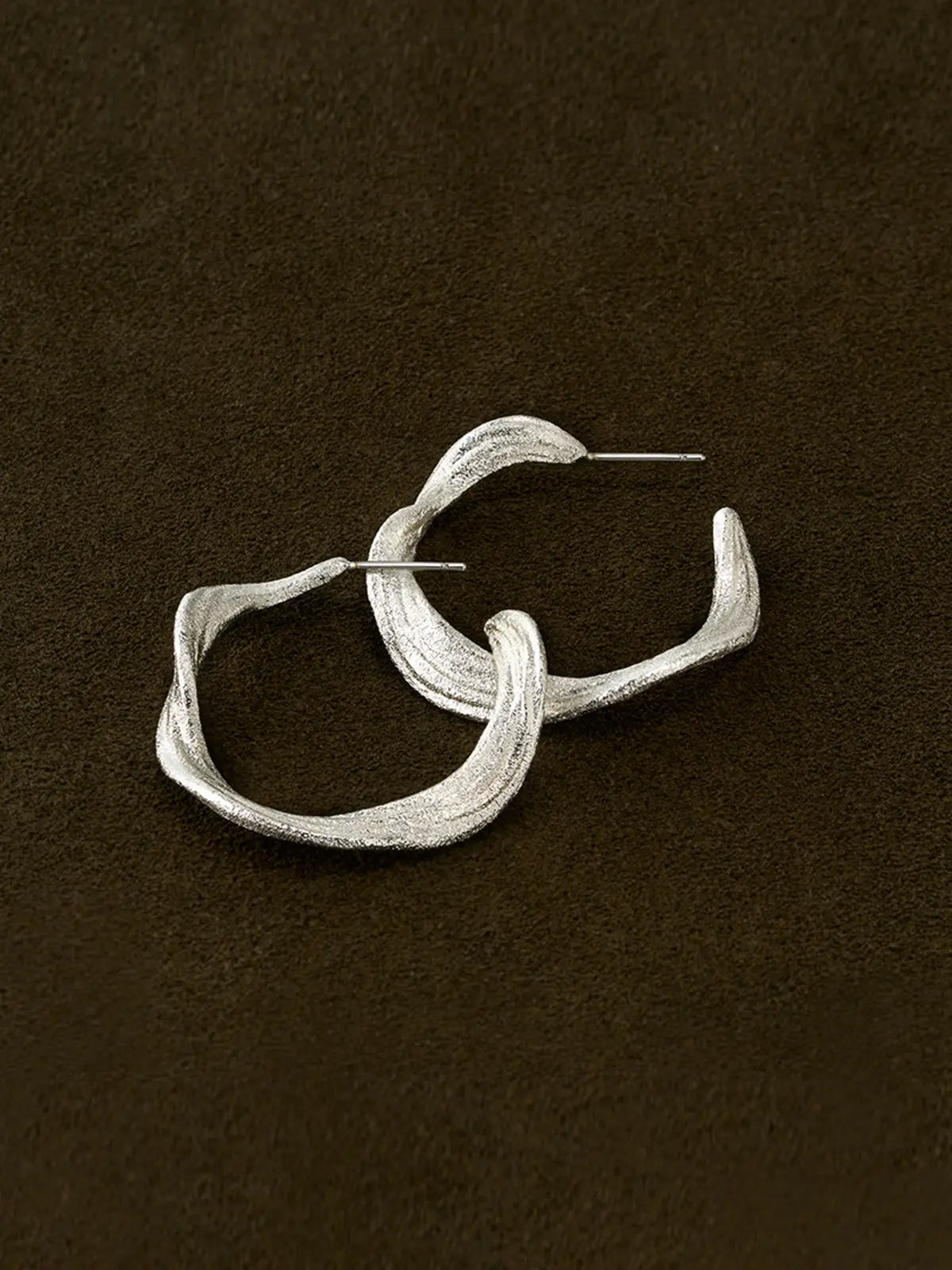 Pair of Tactilery Lake Ripple Hoop Earrings in matte brushed silver displayed lying flat and overlapping on a dark brown suede surface, showing the gently undulating wavy silhouette and fine horizontal brushed ripple texture across both hoops, with silver post pins visible