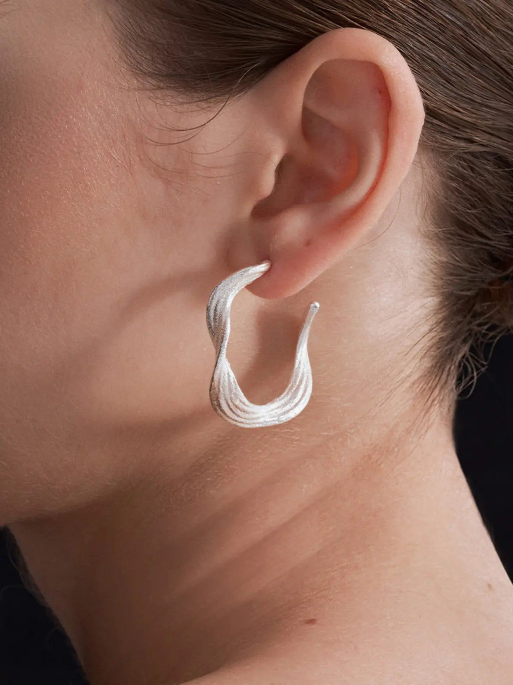 Extreme close-up profile of a model's ear wearing the Tactilery Lake Ripple Hoop Earrings, the large matte brushed silver organic C-hoop with gently undulating wavy silhouette and fine horizontal brushed ripple texture filling the frame, showing the three-dimensional depth of the wide flat band and the open C-hoop construction