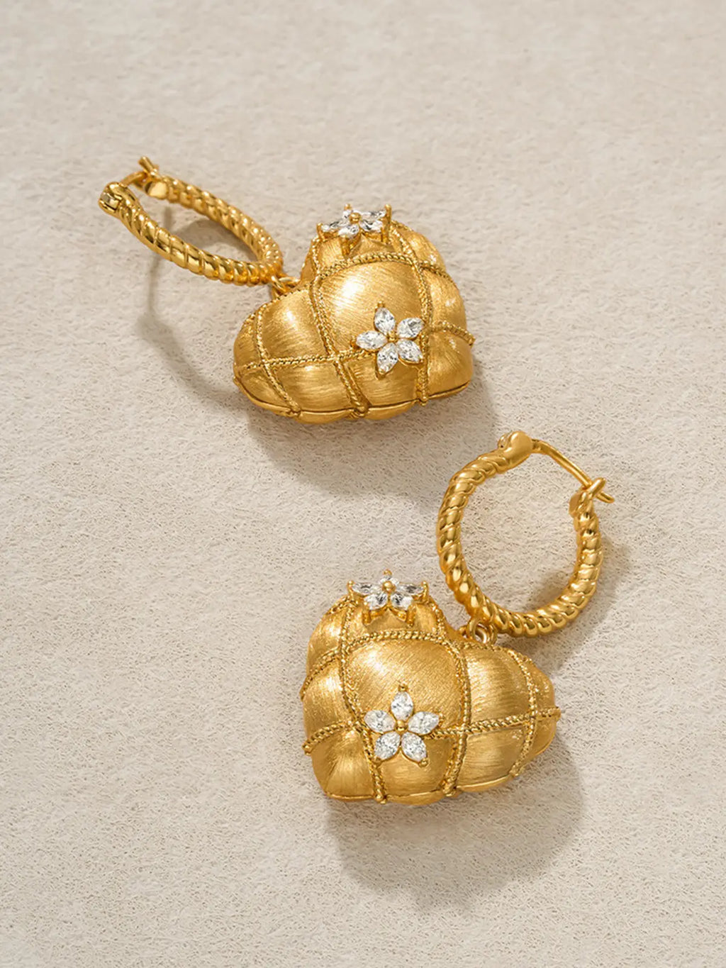 Pair of Tactilery Love Blossom Hoop Earrings displayed at an angle on a warm cream surface, one showing the full front face of the puffy quilted matte gold heart charm with marquise zircon flower clusters and twisted rope hoop, one showing the side profile depth of the three-dimensional heart form