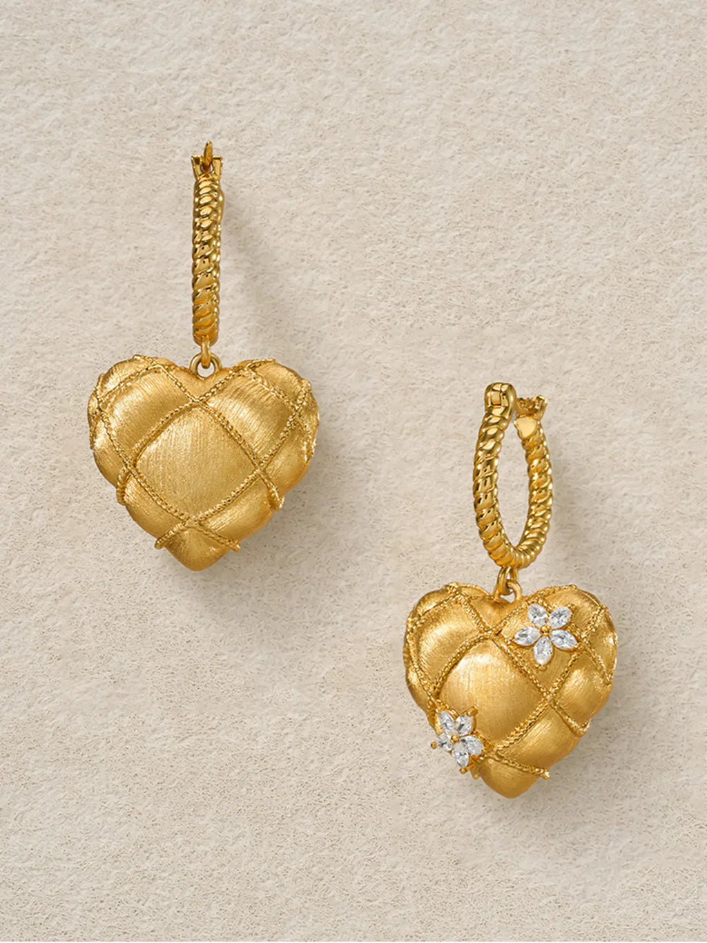 Pair of Tactilery Love Blossom Hoop Earrings displayed face-up on a warm cream surface, showing the full front face of the puffy quilted matte brushed gold heart charm with rope-twist wire grid overlay and two marquise zircon flower clusters, with twisted rope gold hoops visible at top