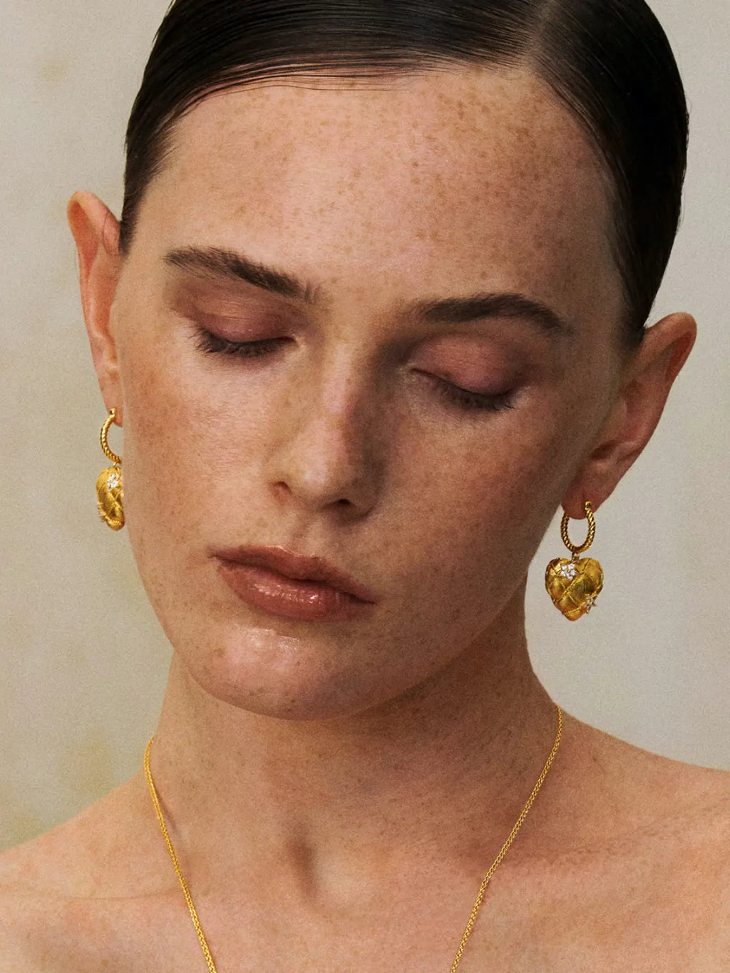 Front-facing editorial portrait of a model wearing the Tactilery Love Blossom Hoop Earrings on both ears, the puffy quilted matte gold heart charm with rope-twist wire grid and marquise zircon flower clusters dangling below the lobe, paired with a delicate gold chain necklace, against a warm beige background with hair slicked back