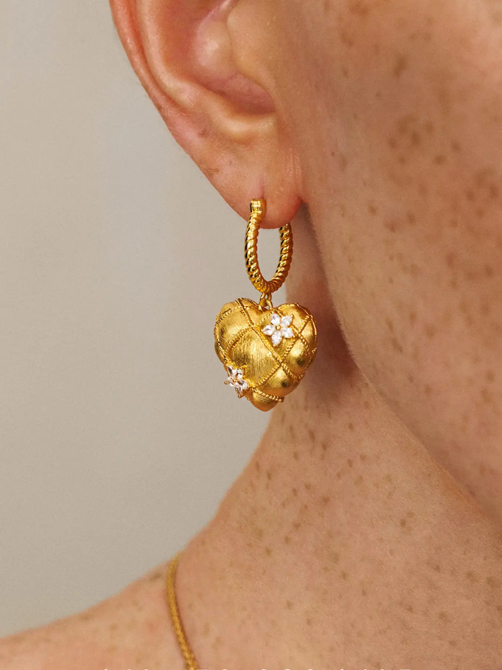 Close-up profile of a model's ear wearing the Tactilery Love Blossom Hoop Earrings, the puffy quilted matte brushed gold heart charm with rope-twist wire grid overlay and two marquise zircon flower clusters dangling from the twisted rope gold hoop, against a warm beige background