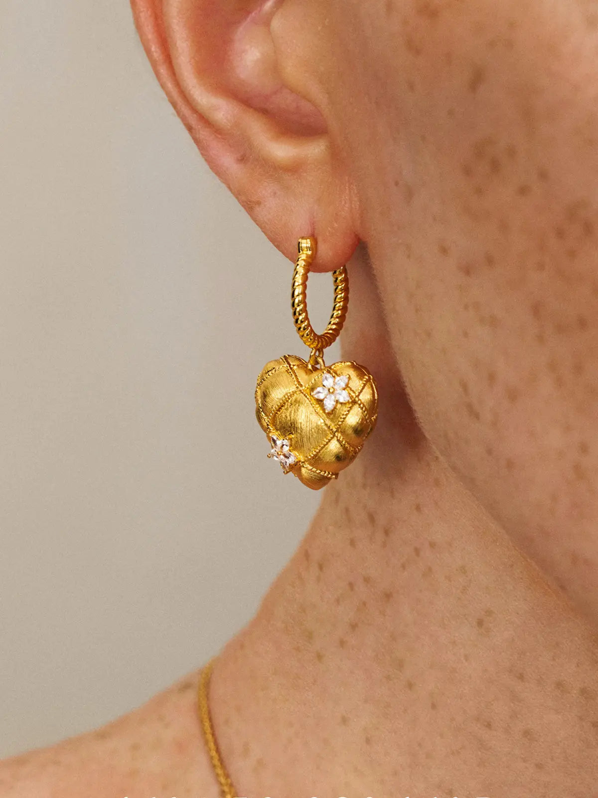 Close-up profile of a model's ear wearing the Tactilery Love Blossom Hoop Earrings, the puffy quilted matte brushed gold heart charm with rope-twist wire grid overlay and two marquise zircon flower clusters dangling from the twisted rope gold hoop, against a warm beige background