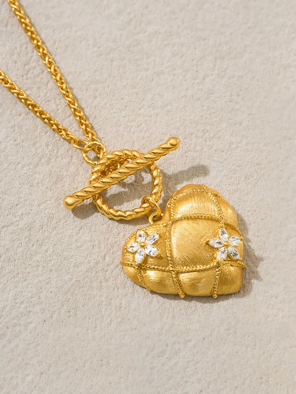 Tactilery Love Blossom Necklace in matte brushed gold displayed lying flat on a warm cream textured surface, the large three-dimensional puffy heart pendant with matte brushed gold quilted diamond-grid surface, fine twisted gold rope milgrain grid borders, and two prong-set four-petal marquise zircon blossom flower accents, suspended from the twisted gold rope OT toggle ring, with the fine polished gold wheat chain and OT toggle bar clasp visible