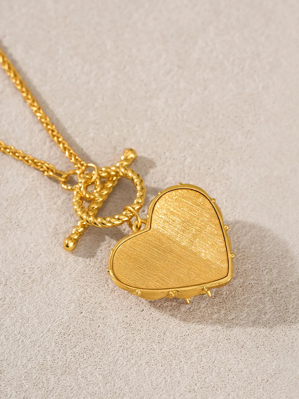 Extreme close-up of the Tactilery Love Blossom Necklace back of pendant on a warm cream textured surface, the large three-dimensional puffy heart pendant shown from the back — the smooth matte brushed gold back face edged by a continuous row of small polished gold ball granulation beads around the perimeter — with the twisted gold rope OT toggle ring and fine polished gold wheat chain visible above