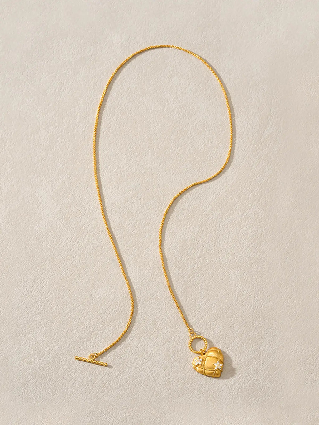 Tactilery Love Blossom Necklace in matte brushed gold displayed laid out flat on a warm cream textured surface showing the full necklace length, the fine polished gold wheat chain extending the full length with the twisted gold rope OT toggle bar clasp at one end and the twisted gold rope OT toggle ring with the large three-dimensional puffy heart pendant at the other end