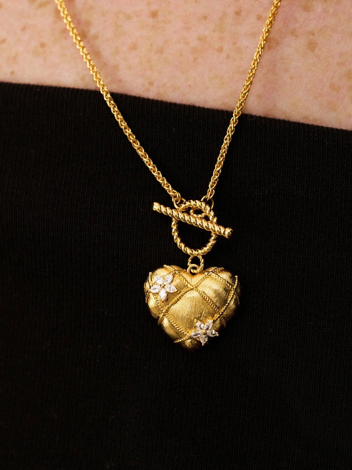 Close-up of a model's décolletage wearing the Tactilery Love Blossom Necklace, the large matte brushed gold quilted diamond-grid puffy heart pendant with two prong-set four-petal marquise zircon blossom flower accents and twisted gold rope OT toggle ring visible at the neckline, on a fine polished gold wheat chain, against a dark background wearing a black top
